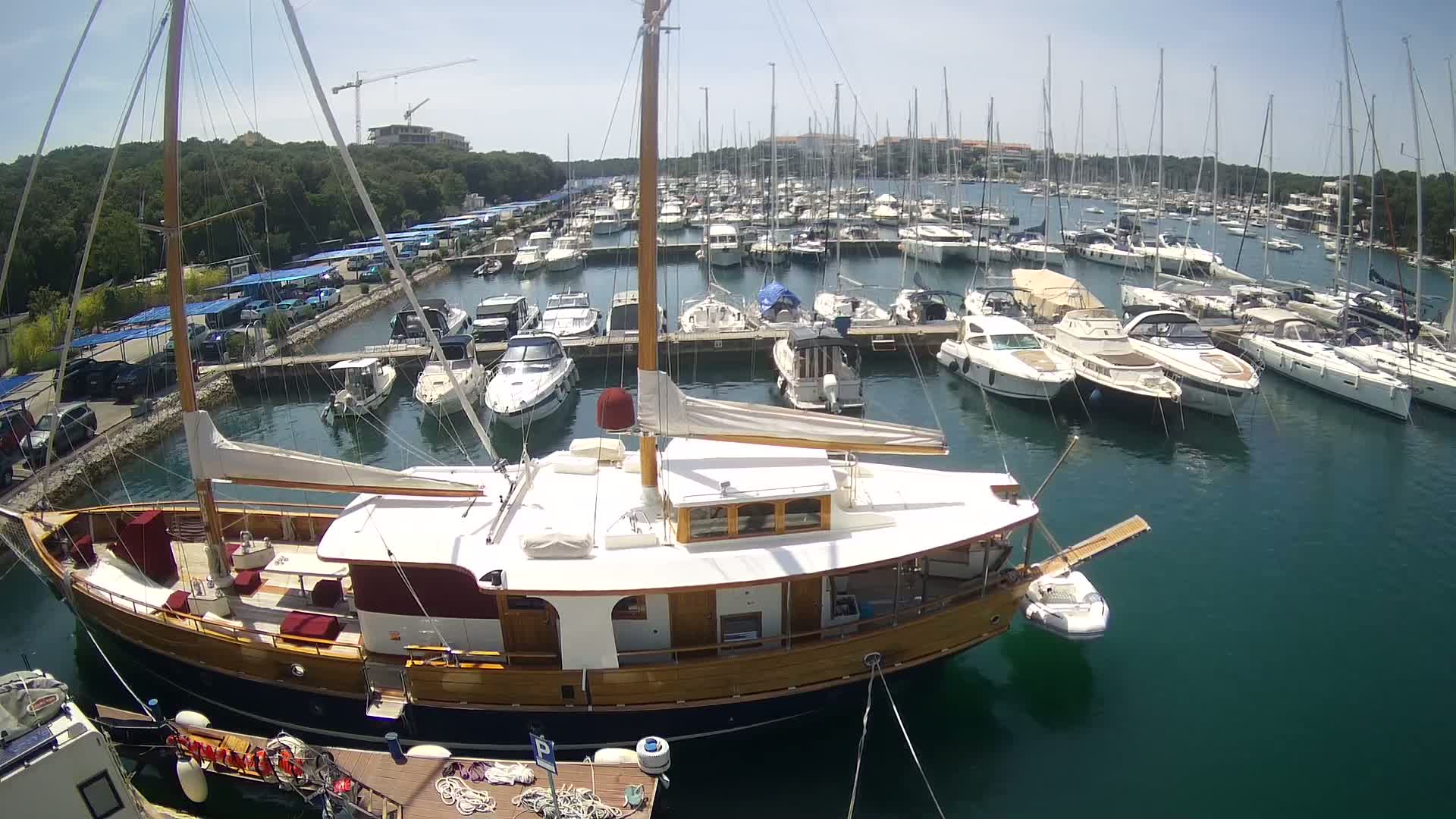 A large wooden sailboat is docked in a marina filled with numerous other sailboats and motorboats on a sunny day.