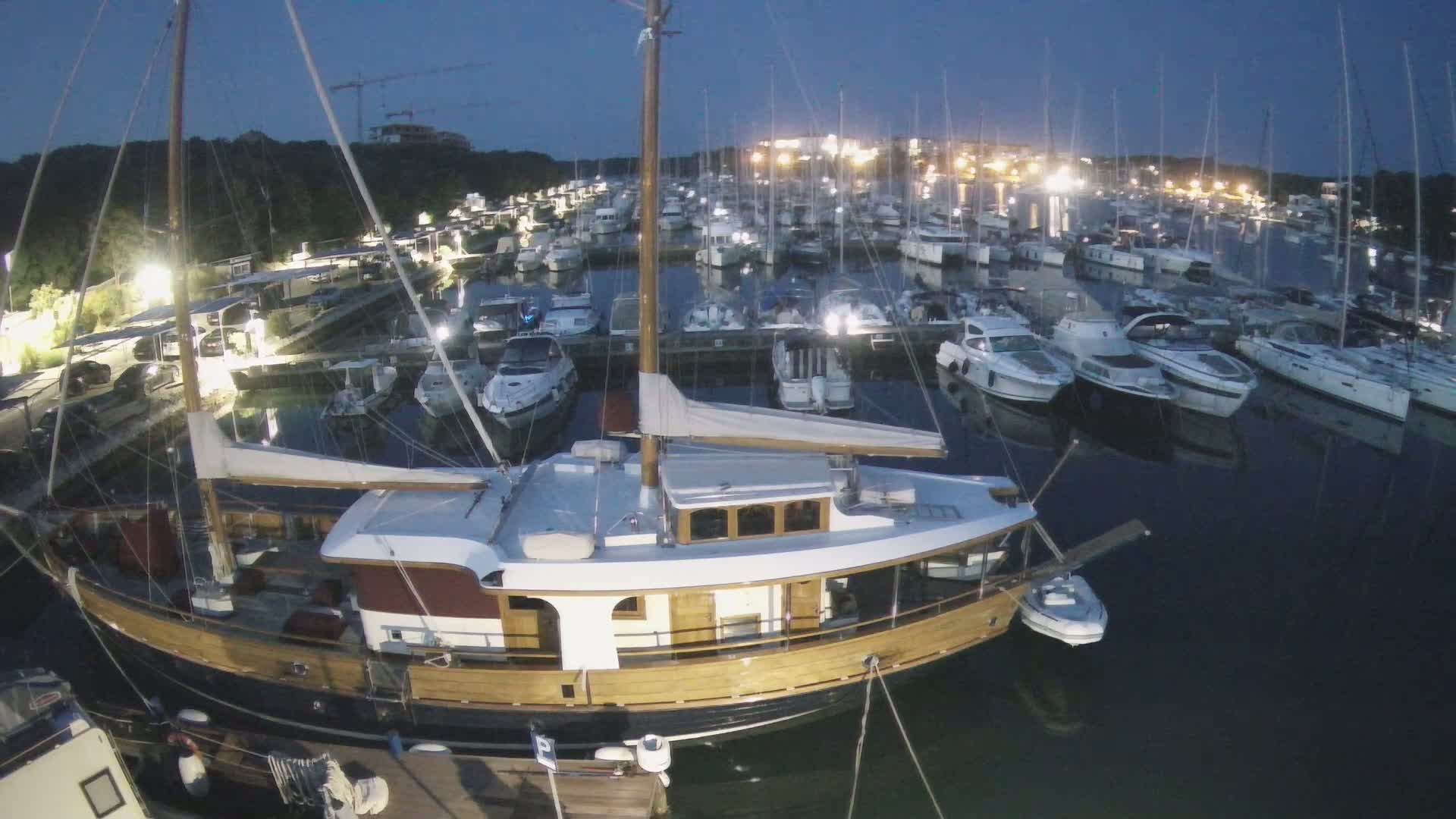 A large wooden sailboat is moored in a marina filled with numerous other boats at night under a dark sky.