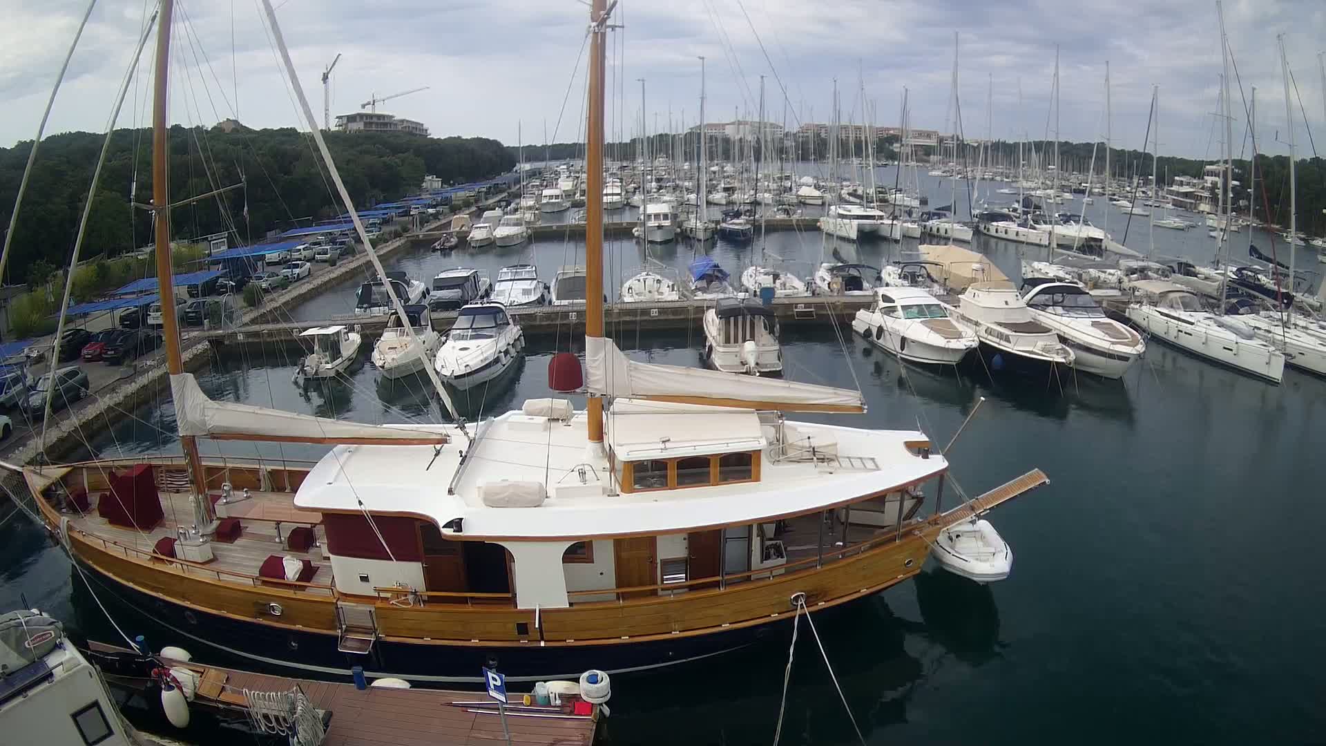 A large wooden sailboat is moored in a marina filled with numerous other sailboats and motorboats under a cloudy sky.