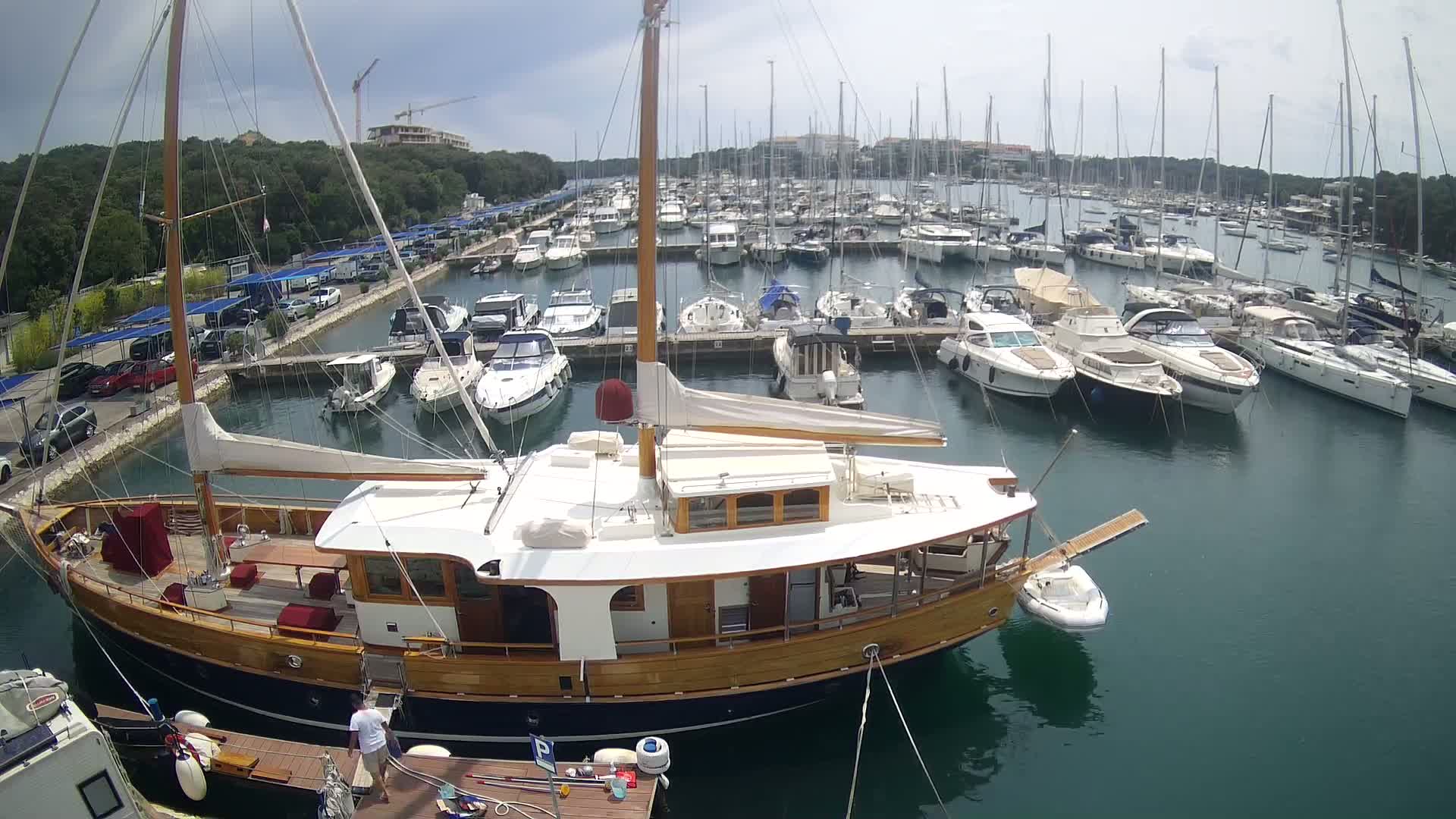 A large wooden sailboat is docked in a marina filled with numerous other boats on a partly cloudy day.