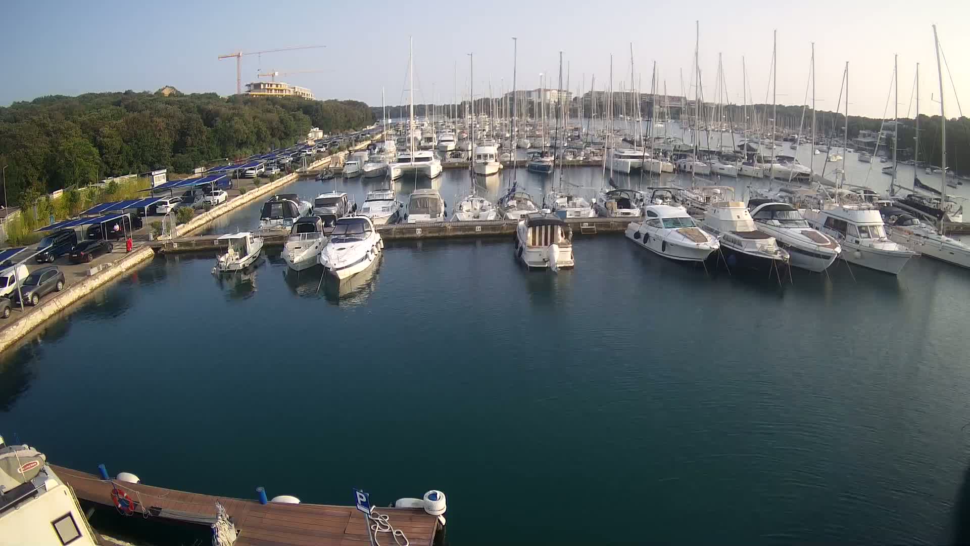 A marina filled with numerous boats and yachts on a calm, sunny day, with a parking lot and trees visible in the background.
