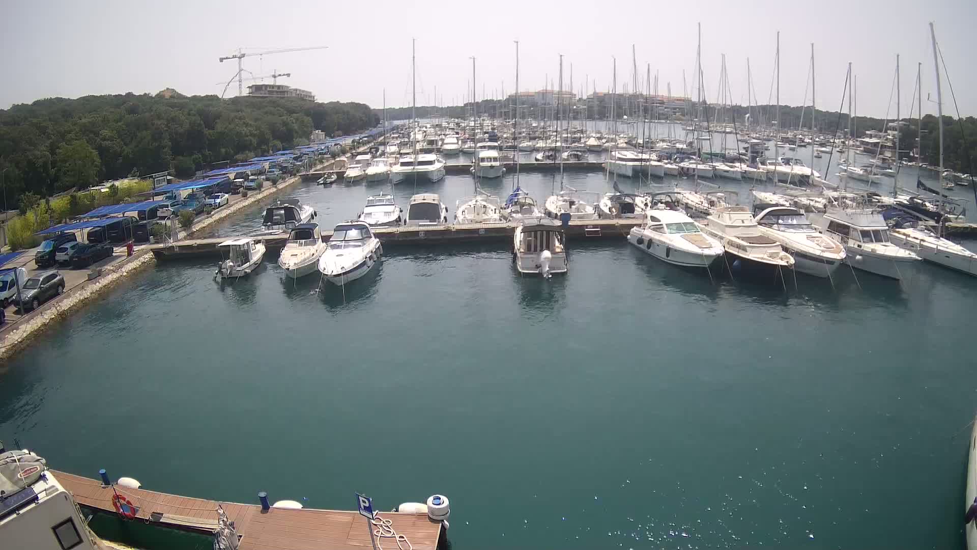 A marina filled with numerous boats and yachts on a sunny day, with cars parked alongside a dock in the foreground.