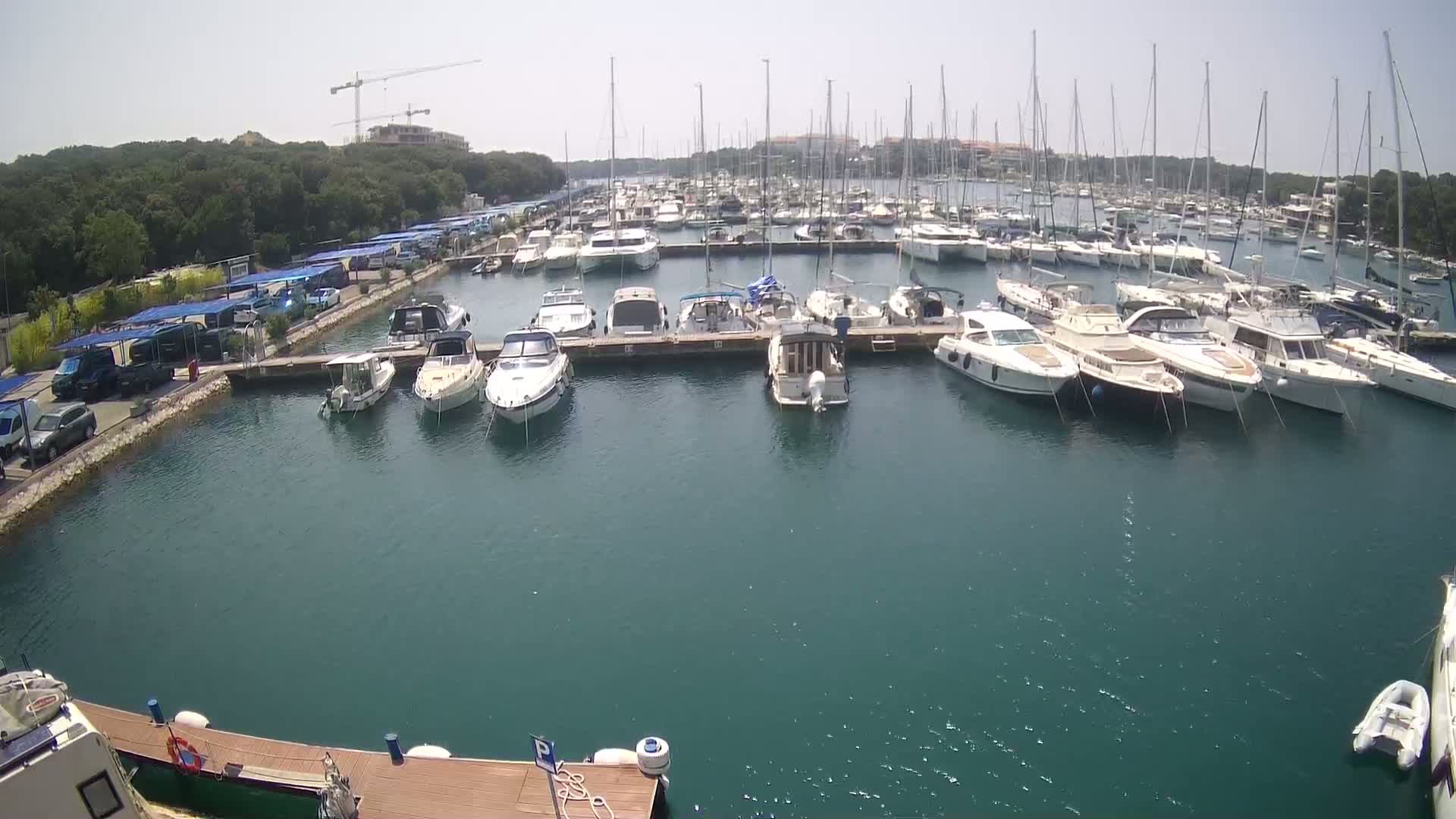 A marina filled with numerous yachts and motorboats is shown on a sunny day, with a parking lot and green trees visible in the background.