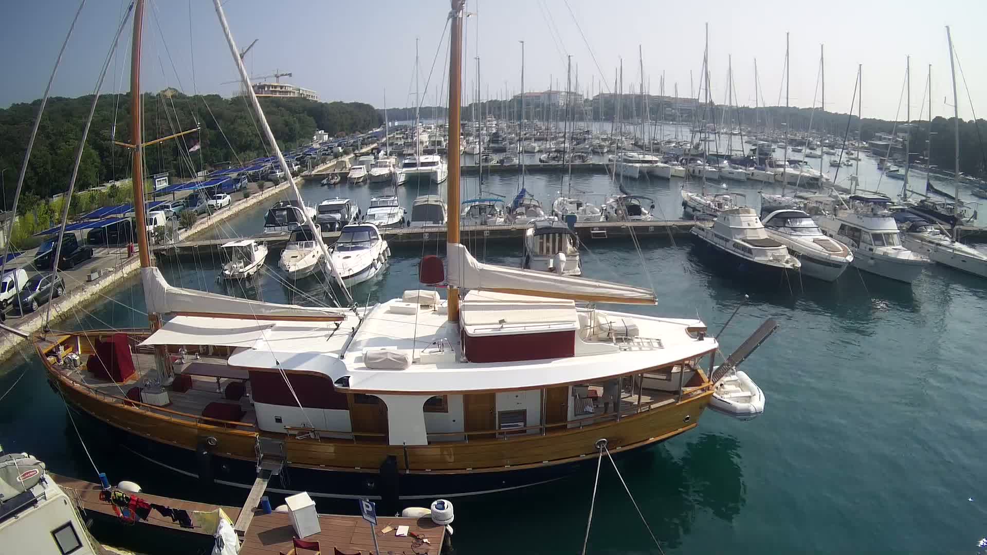 A large wooden sailboat is docked in a marina filled with numerous other boats on a sunny day.