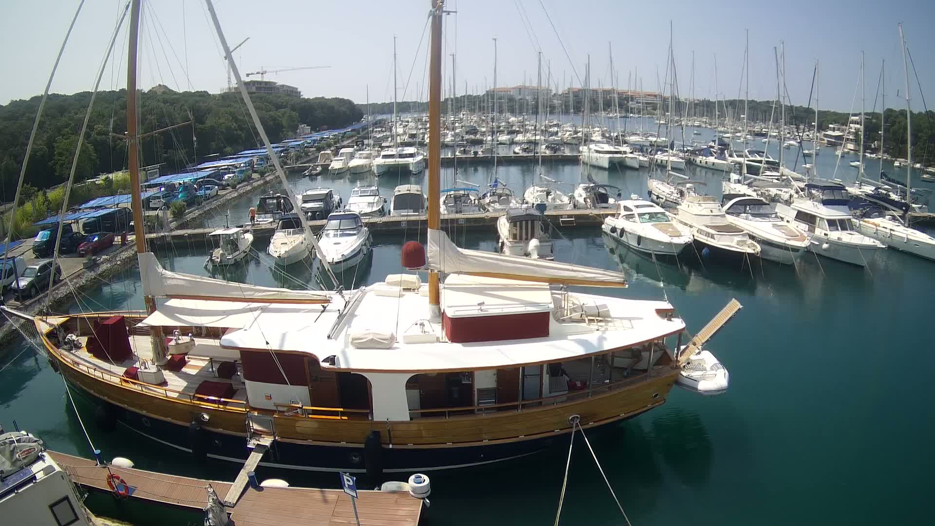 A large wooden sailboat is docked in a marina filled with numerous other boats on a sunny day.