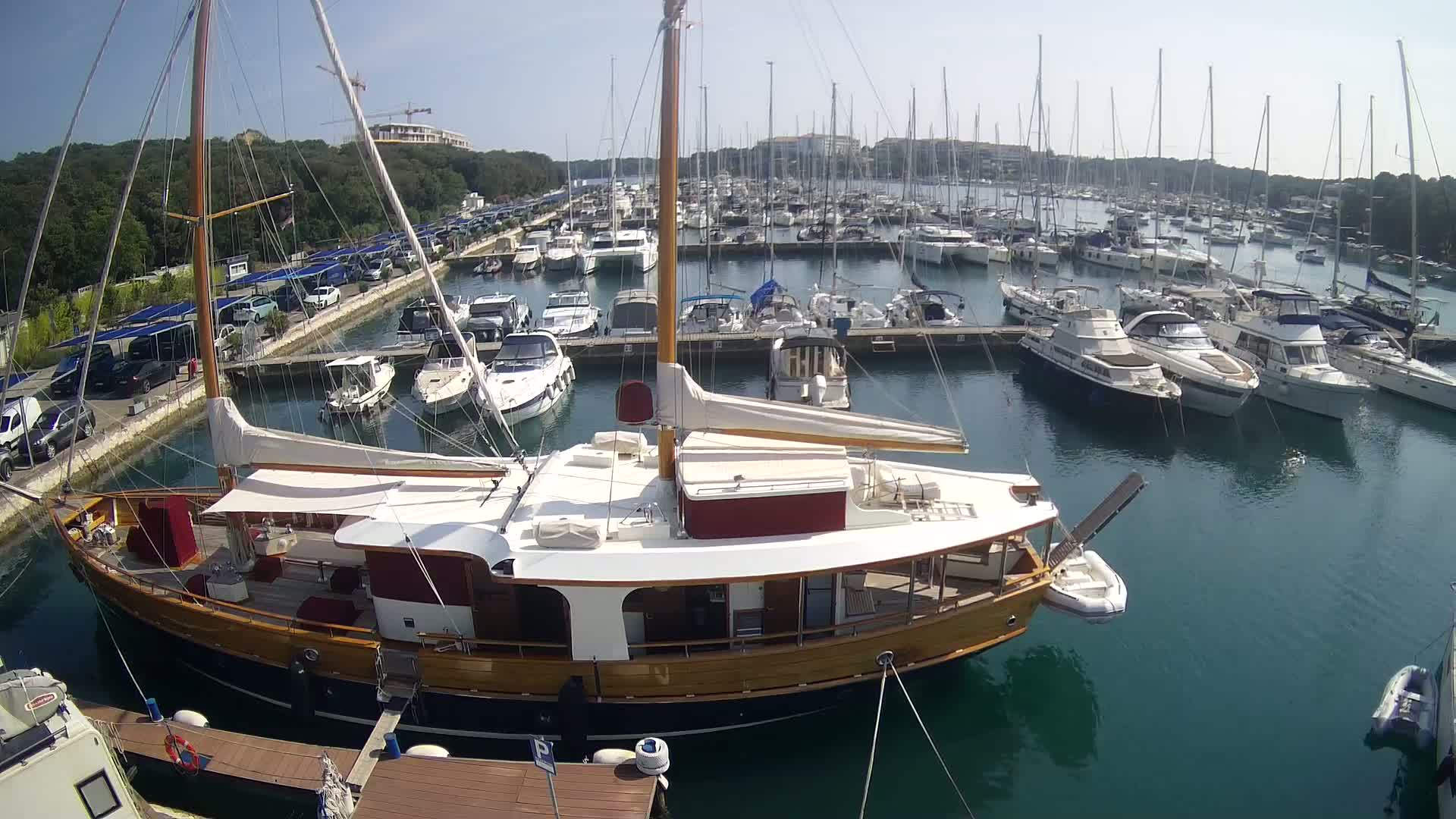 A large wooden sailboat is docked in a marina filled with numerous other boats on a sunny day.
