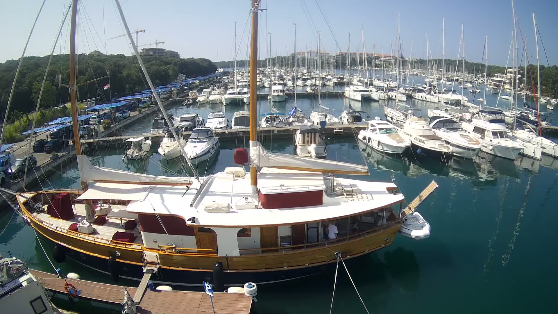 A large wooden sailboat is docked in a marina filled with numerous other boats on a sunny day.