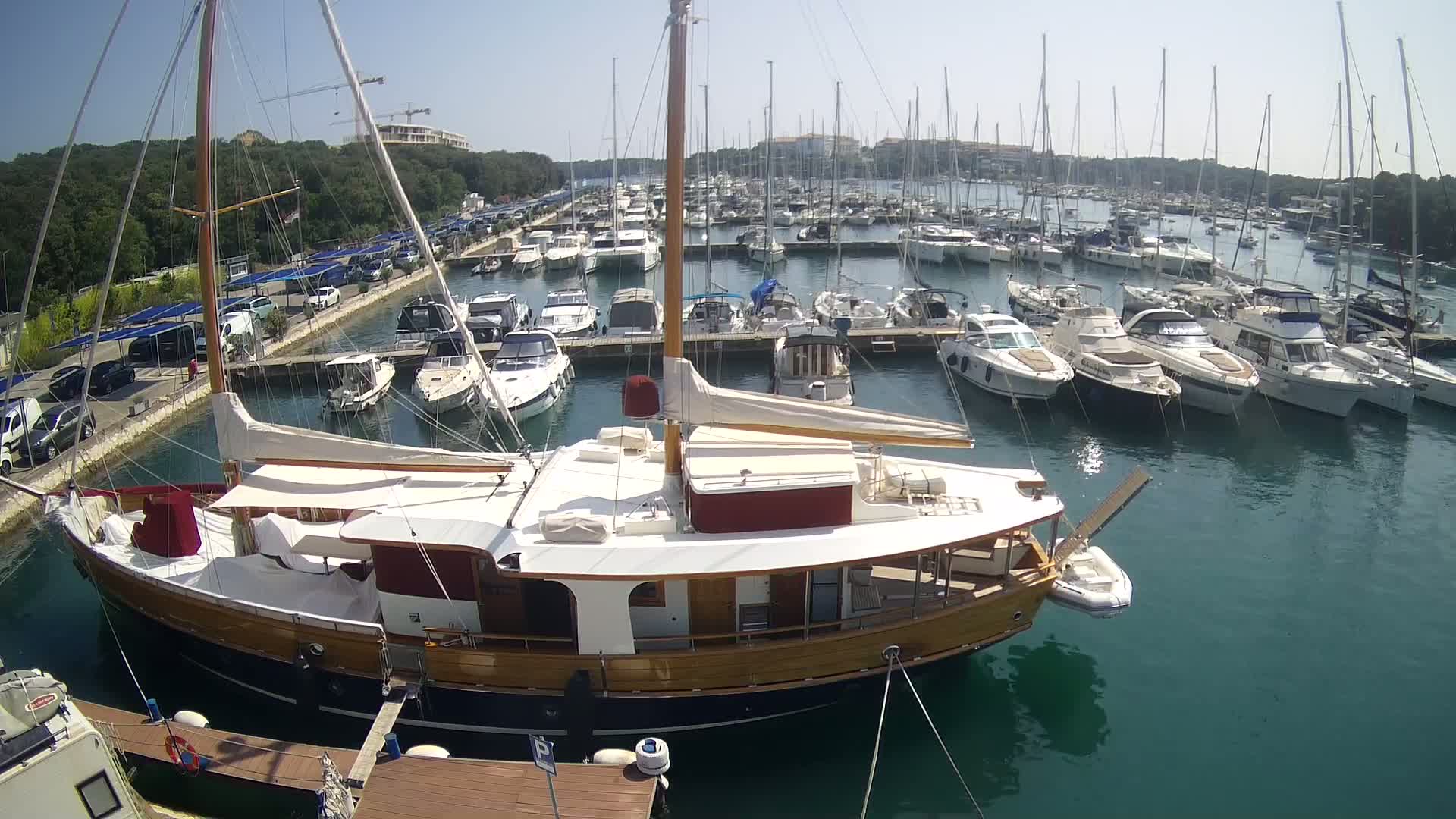 A large wooden sailboat is docked in a marina filled with numerous other boats on a sunny day.