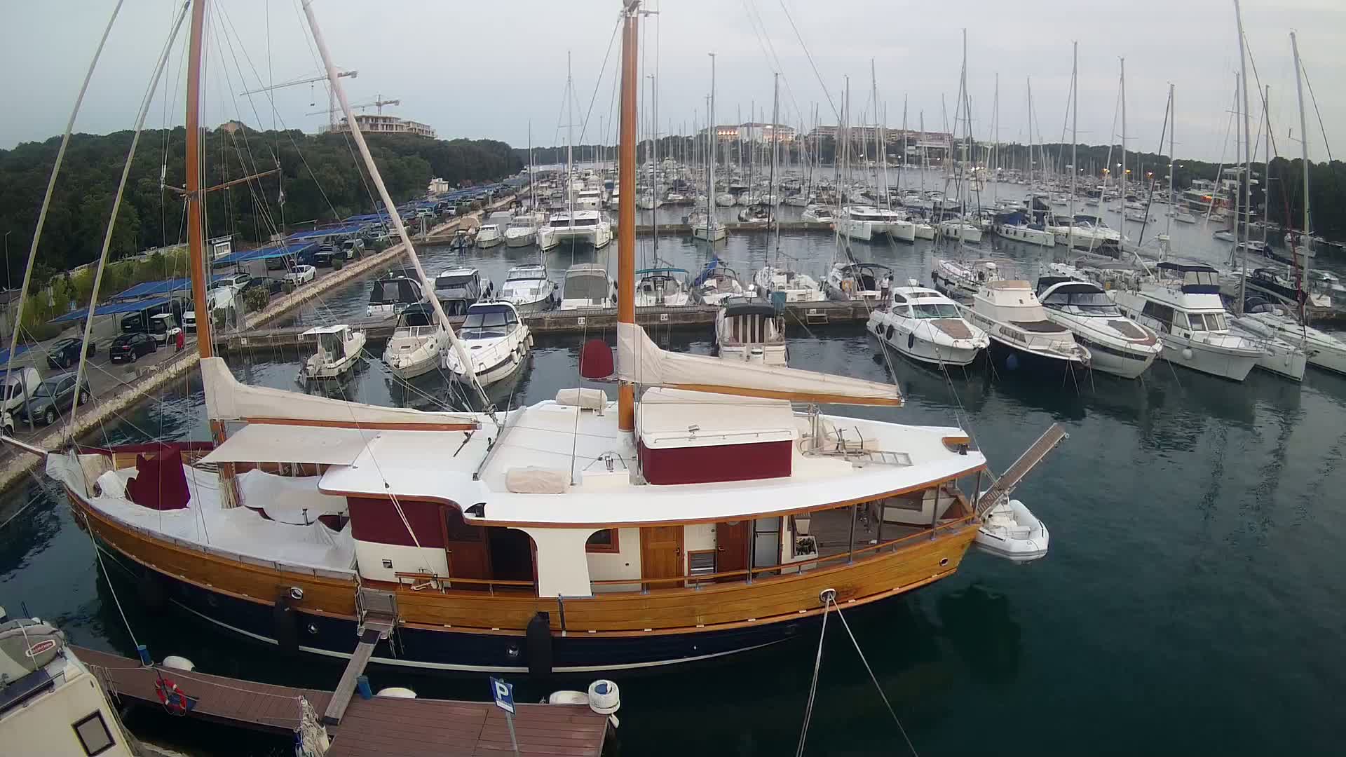 A large wooden sailboat is docked in a marina filled with numerous other boats on a sunny day.