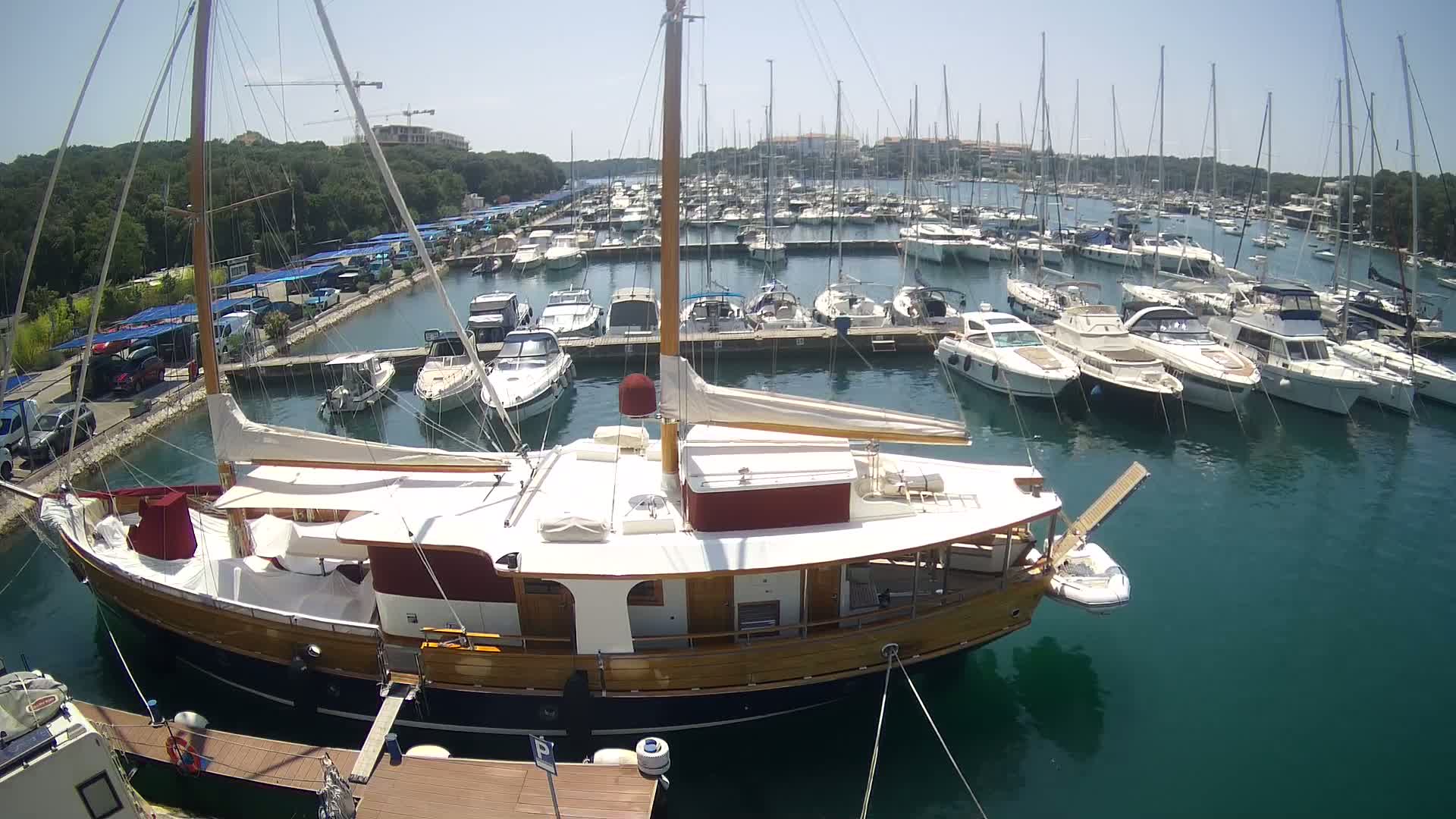 A large wooden sailboat is docked in a marina filled with numerous other boats on a sunny day.