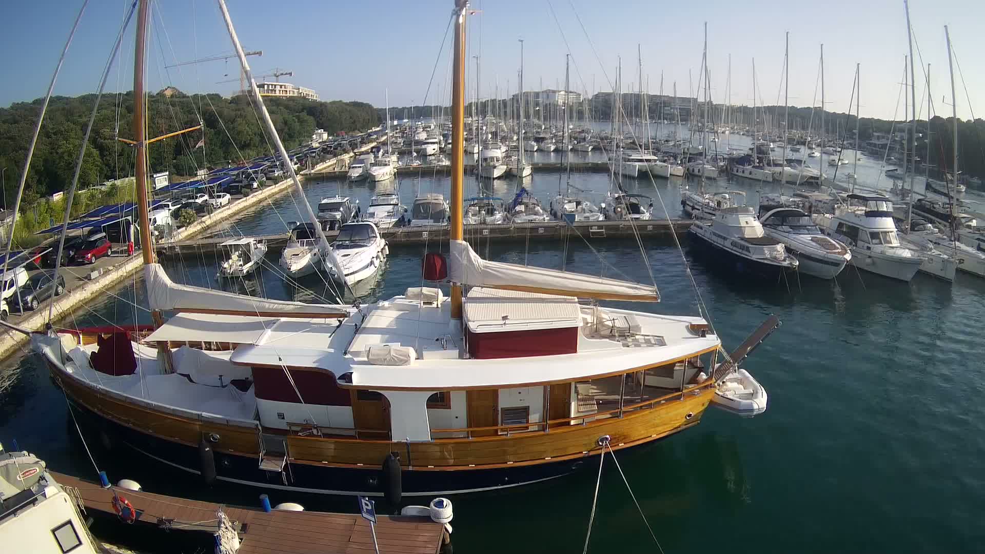 A large wooden sailboat is docked in a marina filled with numerous other boats on a sunny day.