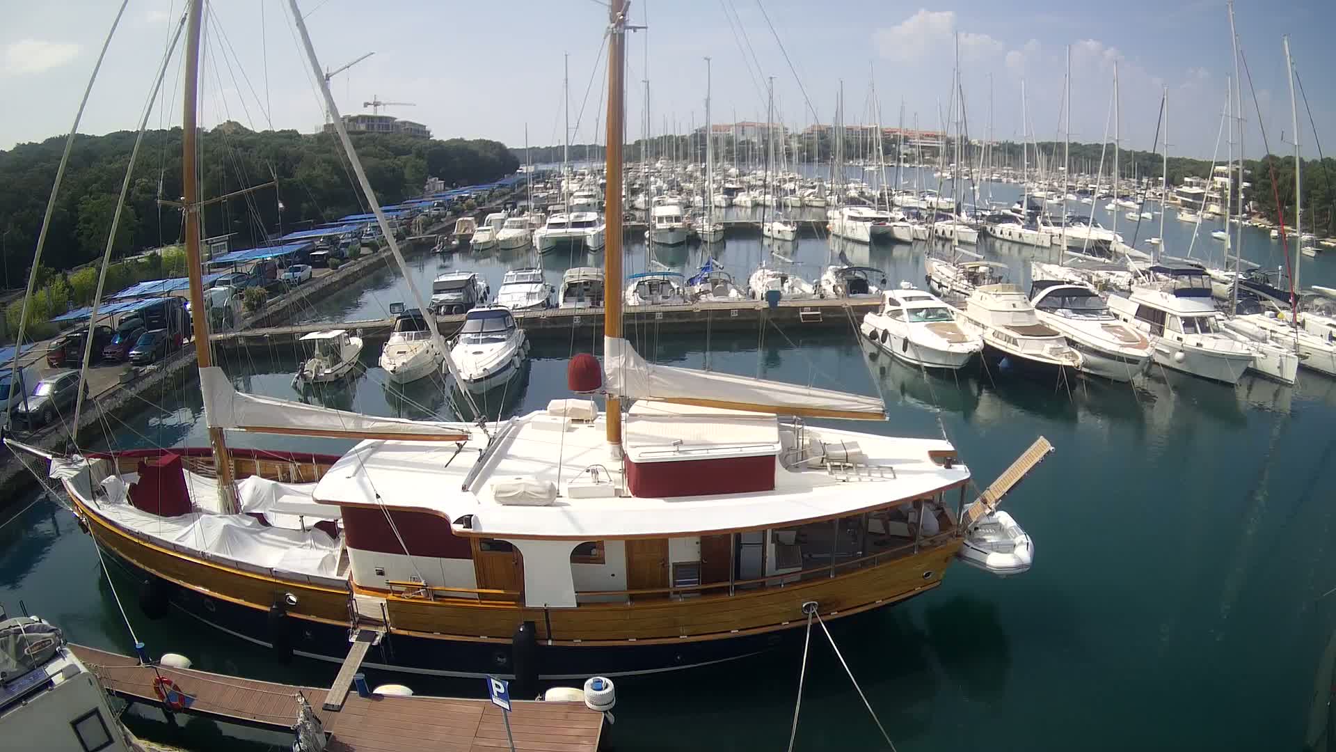 A large wooden sailboat is docked in a marina filled with numerous other boats on a sunny day.