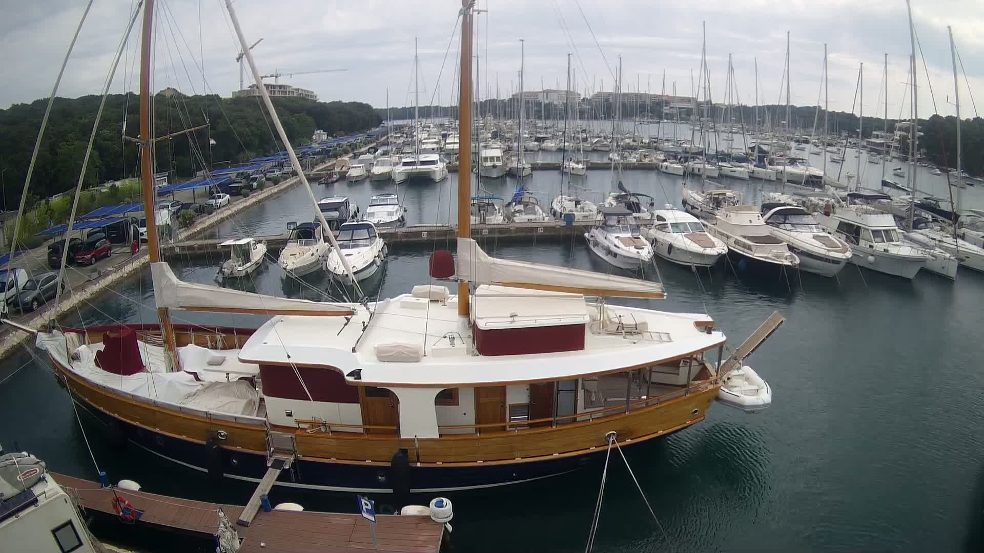 A large wooden sailboat is moored in a marina filled with numerous other sailboats and motorboats under an overcast sky.