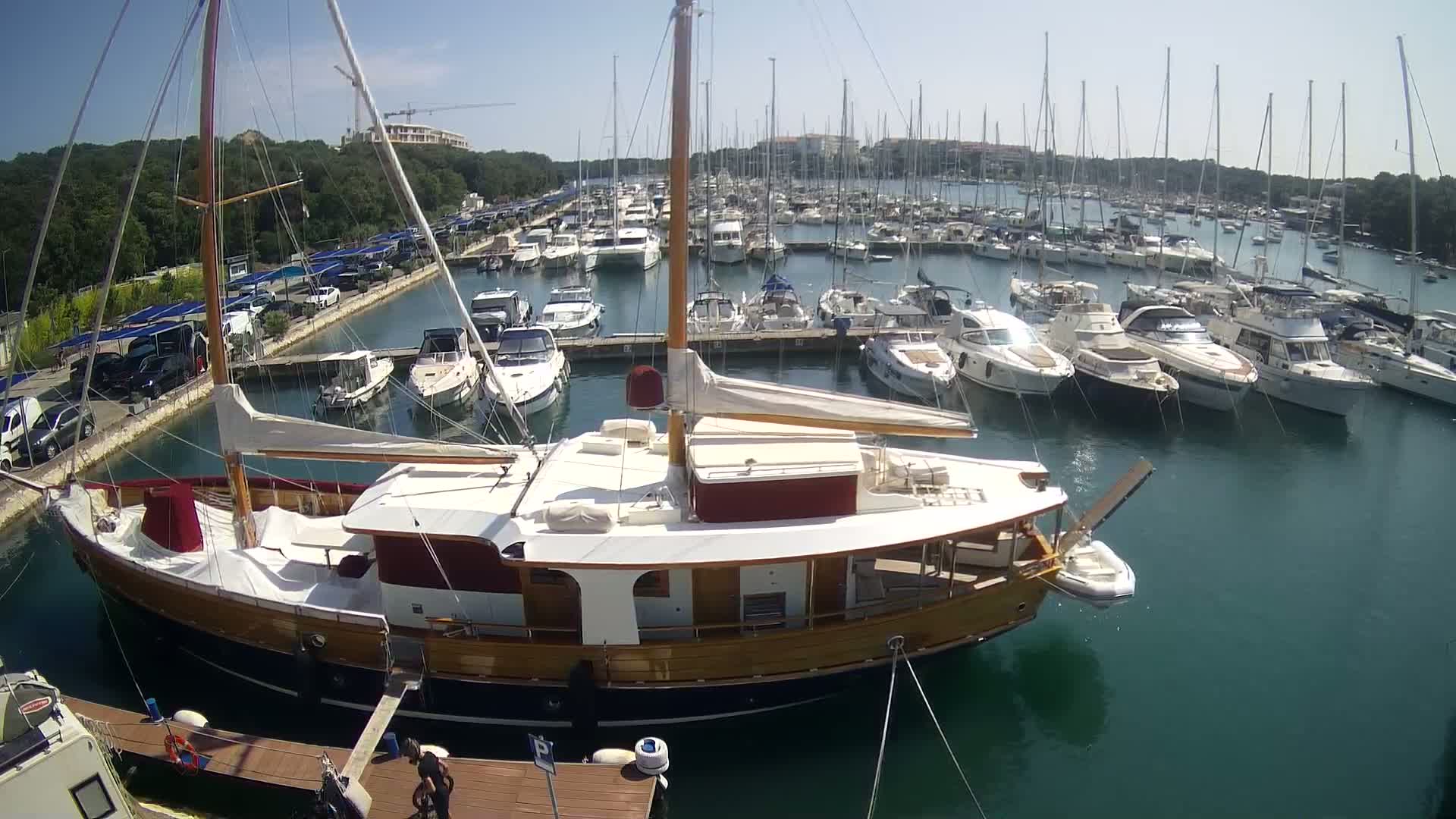 A large wooden sailboat is docked in a marina filled with numerous other boats on a sunny day.