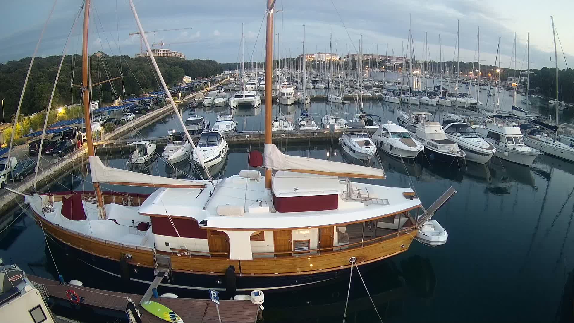 A large wooden sailboat is docked in a marina filled with numerous other boats under a partly cloudy evening sky.