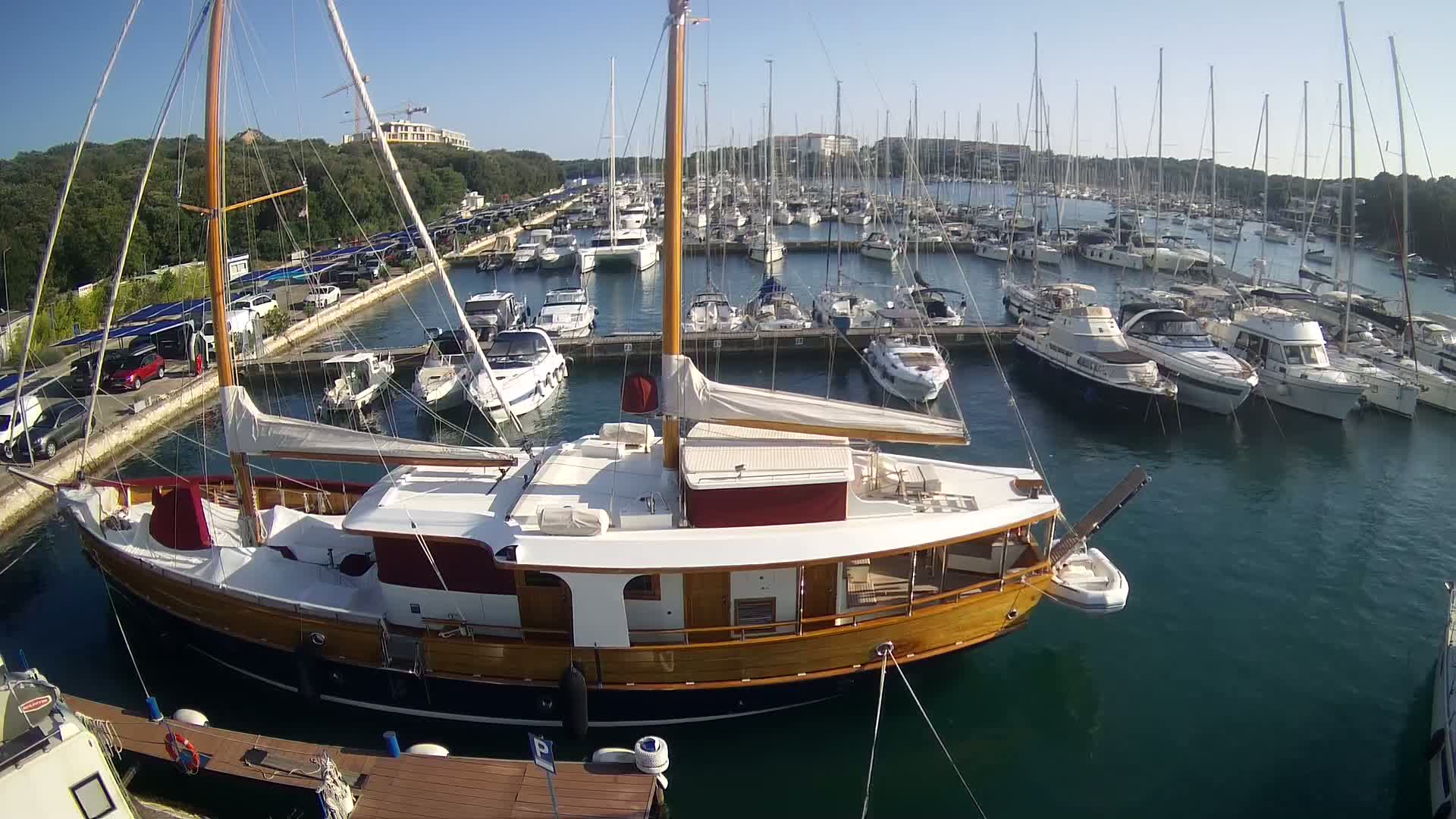 A large, wooden sailboat is docked in a marina filled with numerous other boats on a sunny day.