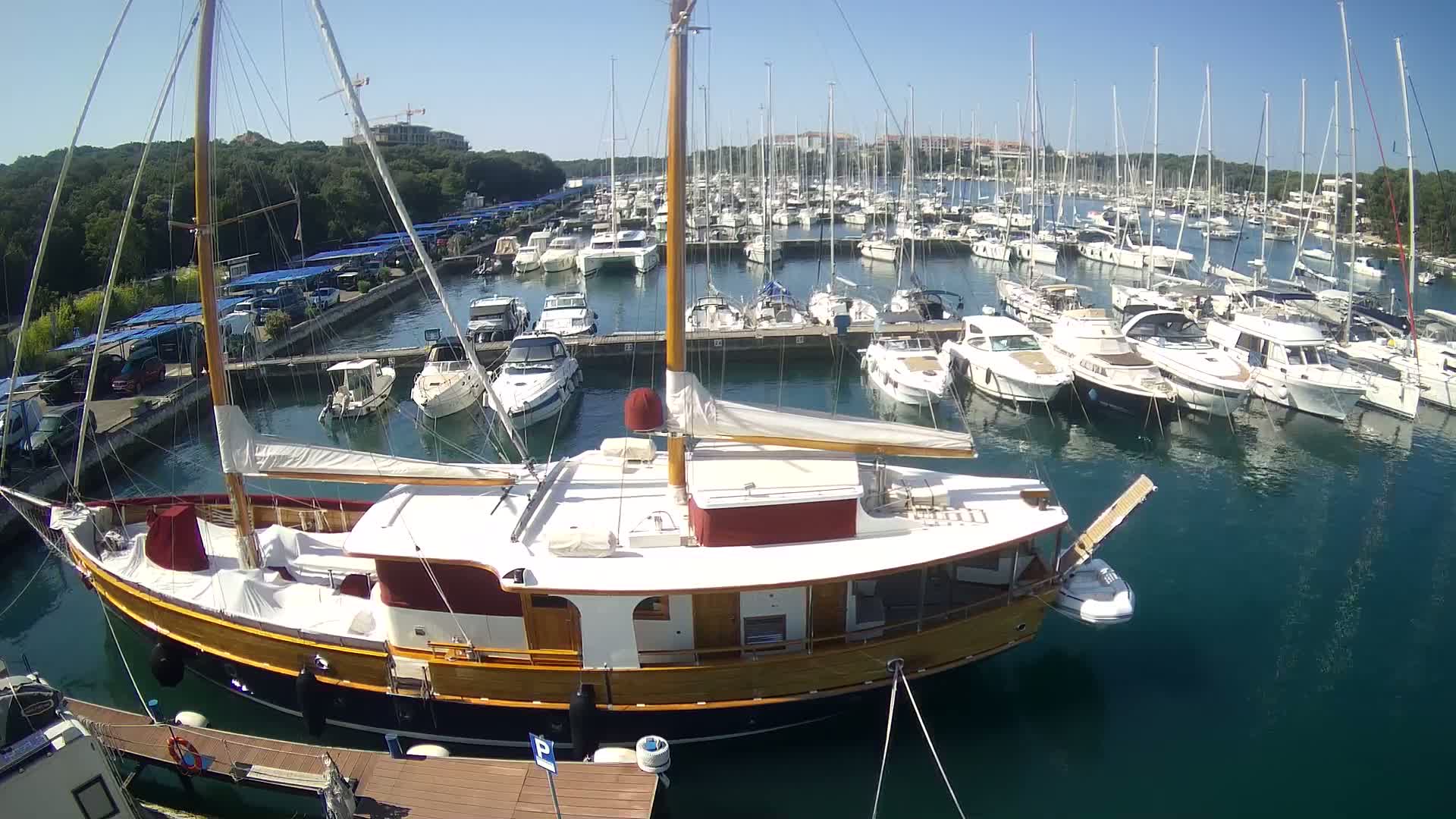 A large wooden sailboat is docked in a marina filled with numerous other sailboats and motorboats on a sunny day.