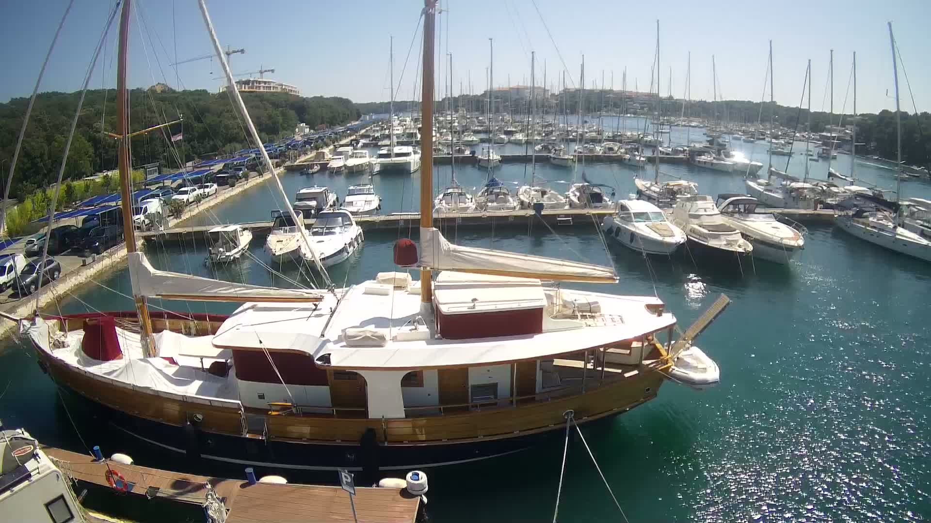 A large wooden sailboat is docked in a marina filled with numerous other boats on a sunny day.