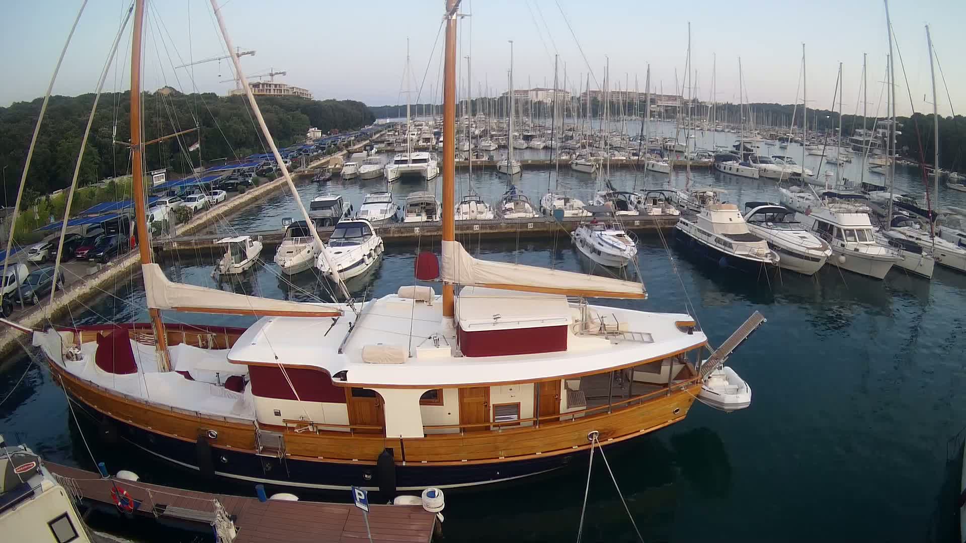 A large wooden sailboat is docked in a marina filled with numerous other boats on a calm, clear day.