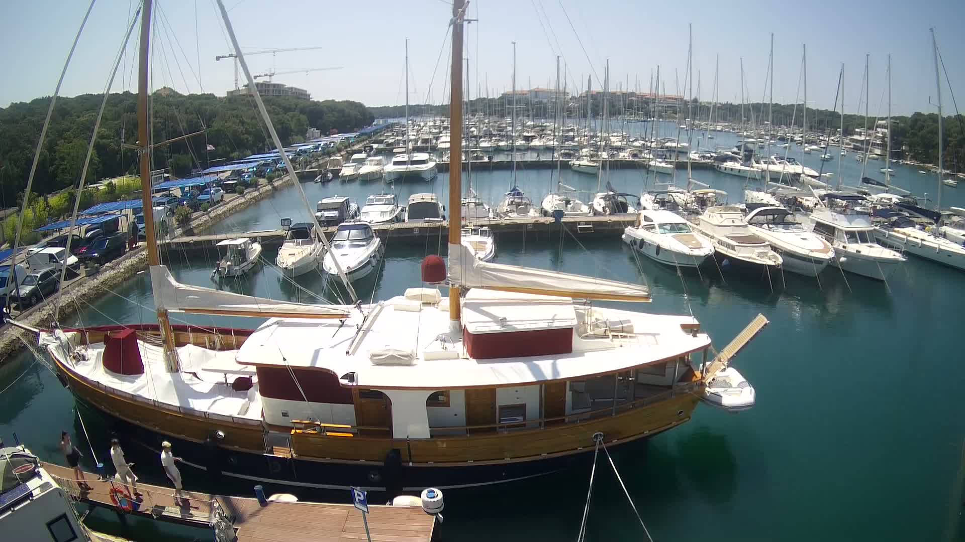 A large wooden sailboat is docked in a marina filled with numerous other boats on a sunny day.