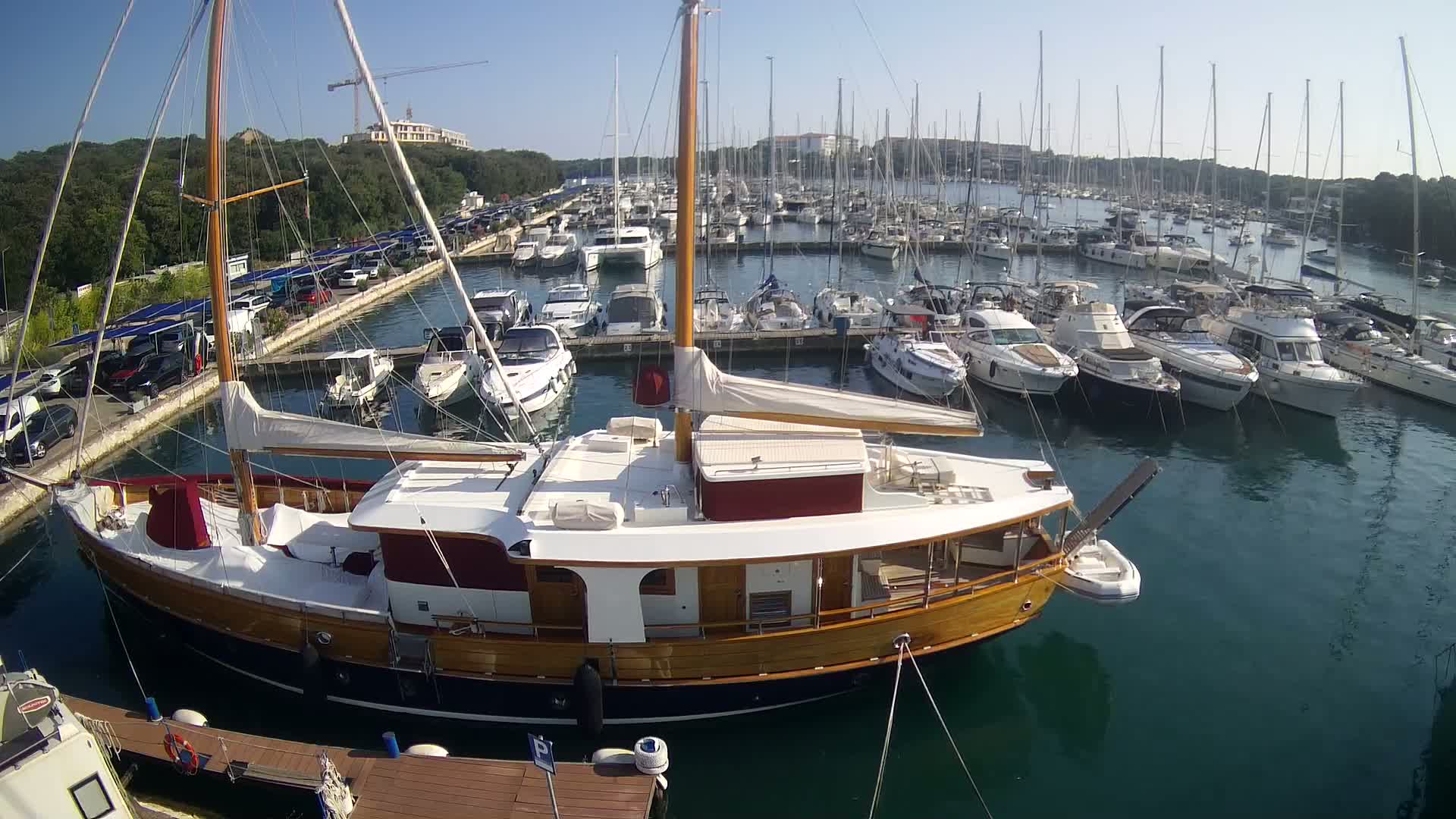 A large wooden sailboat is docked in a marina filled with numerous other boats on a sunny day.