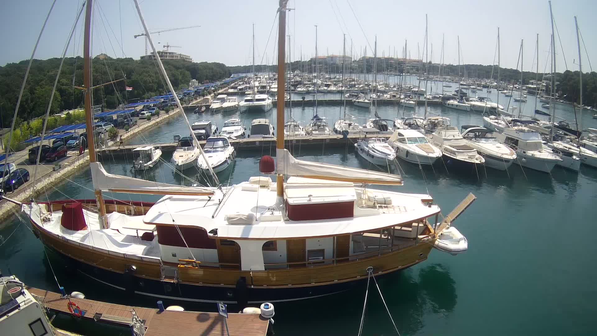 A large wooden sailboat is moored in a marina filled with numerous other sailboats and motorboats on a sunny day.