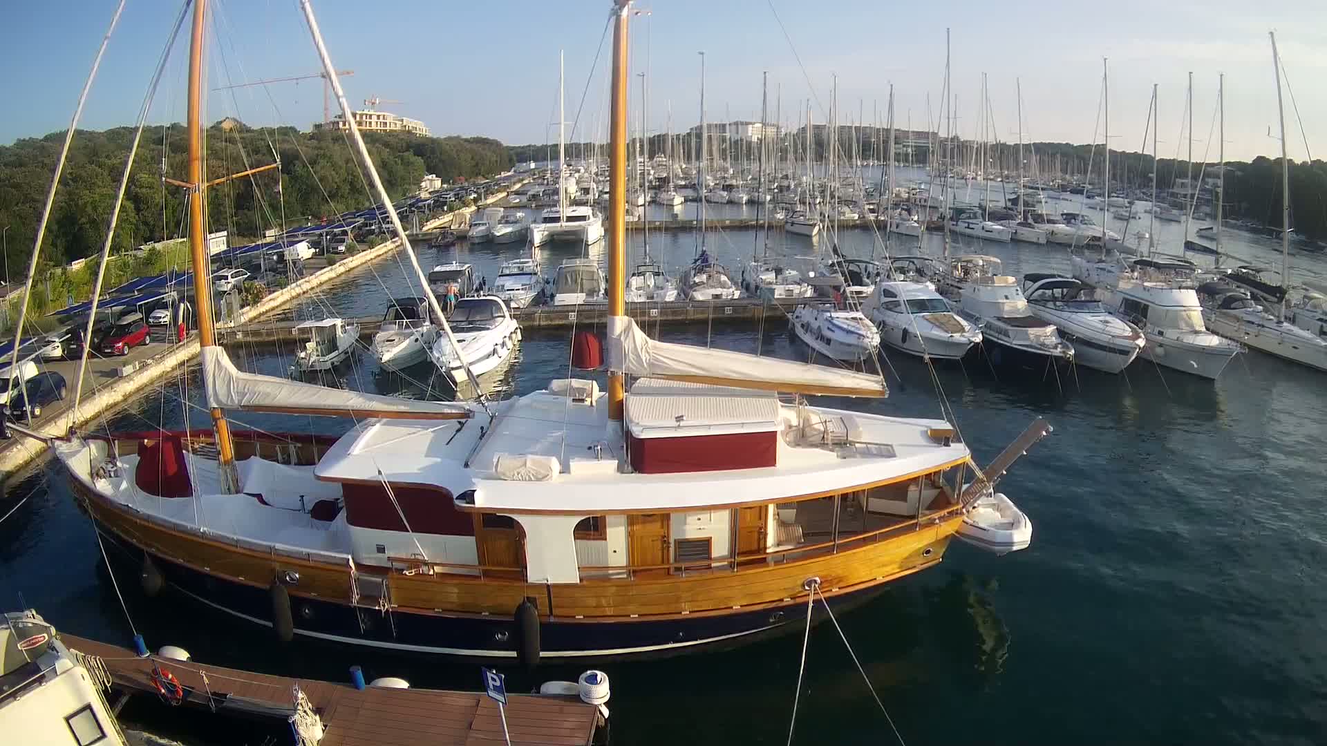 A large wooden sailboat is docked in a marina filled with numerous other boats on a sunny day.