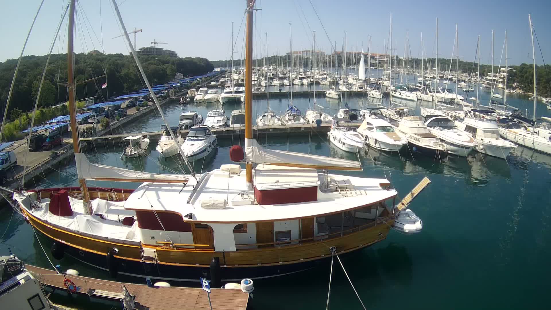 A large wooden sailboat is docked in a marina filled with numerous other boats on a sunny day.