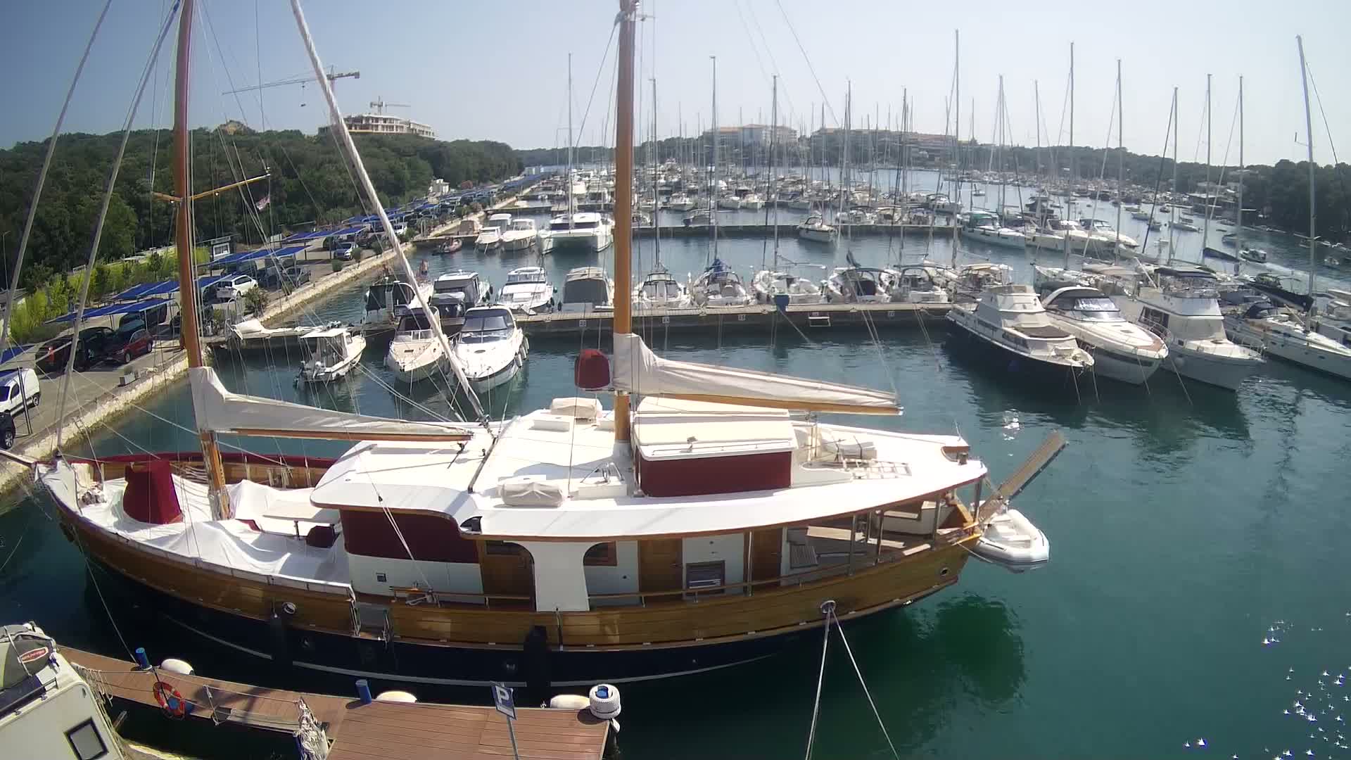 A large wooden sailboat is docked in a marina filled with numerous other boats on a sunny day.