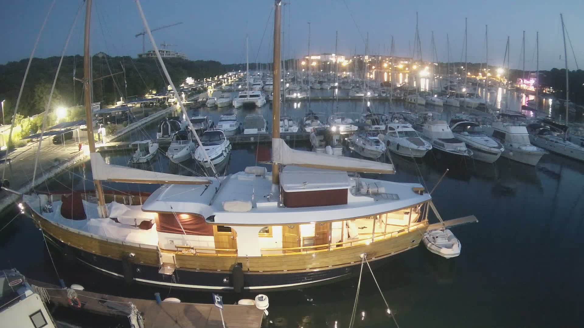 A large wooden sailboat is docked in a marina at night, surrounded by numerous other boats under a twilight sky.