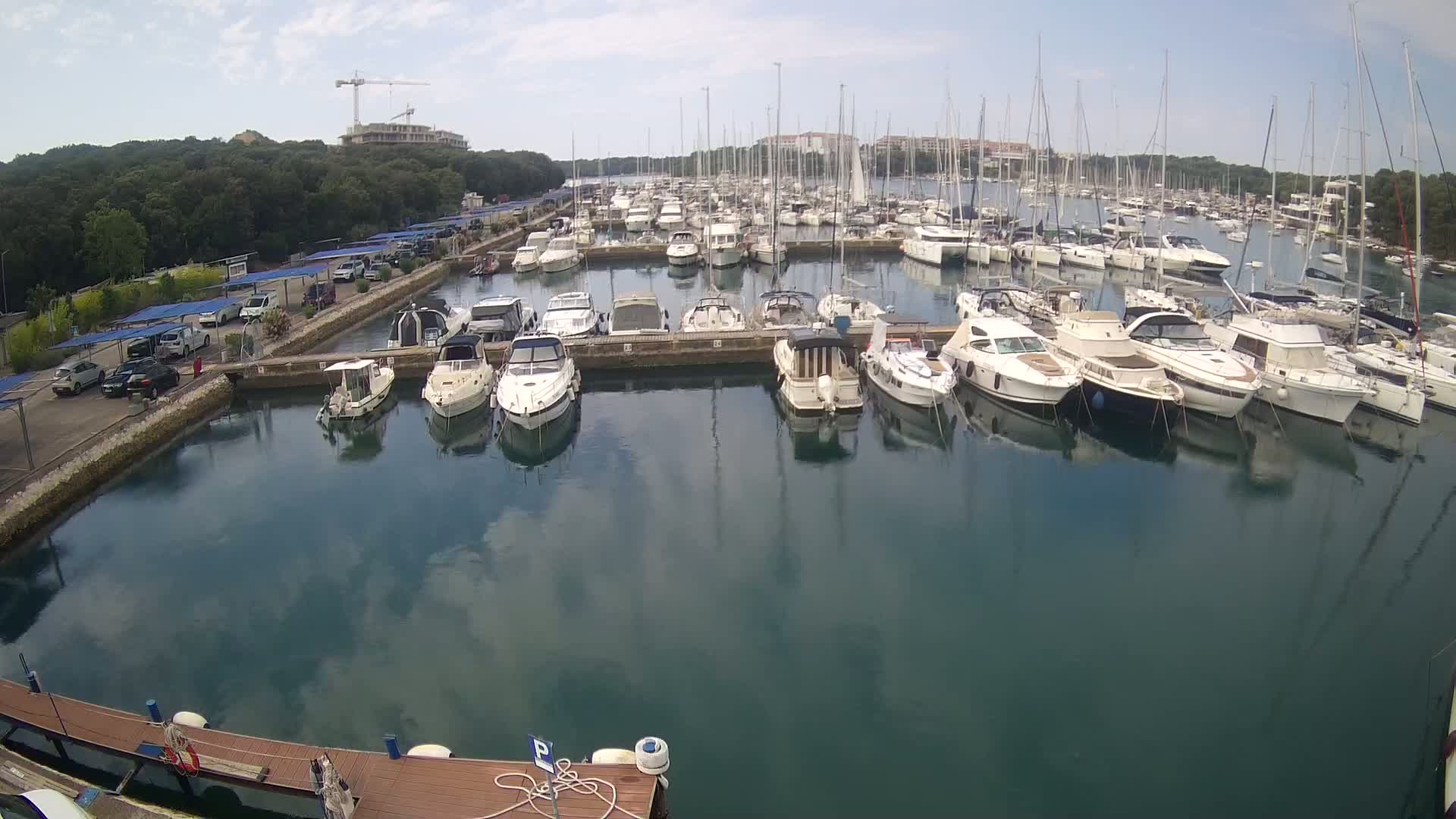Many boats and yachts are docked in a calm marina on a partly sunny day, with a parking lot and trees visible in the background.