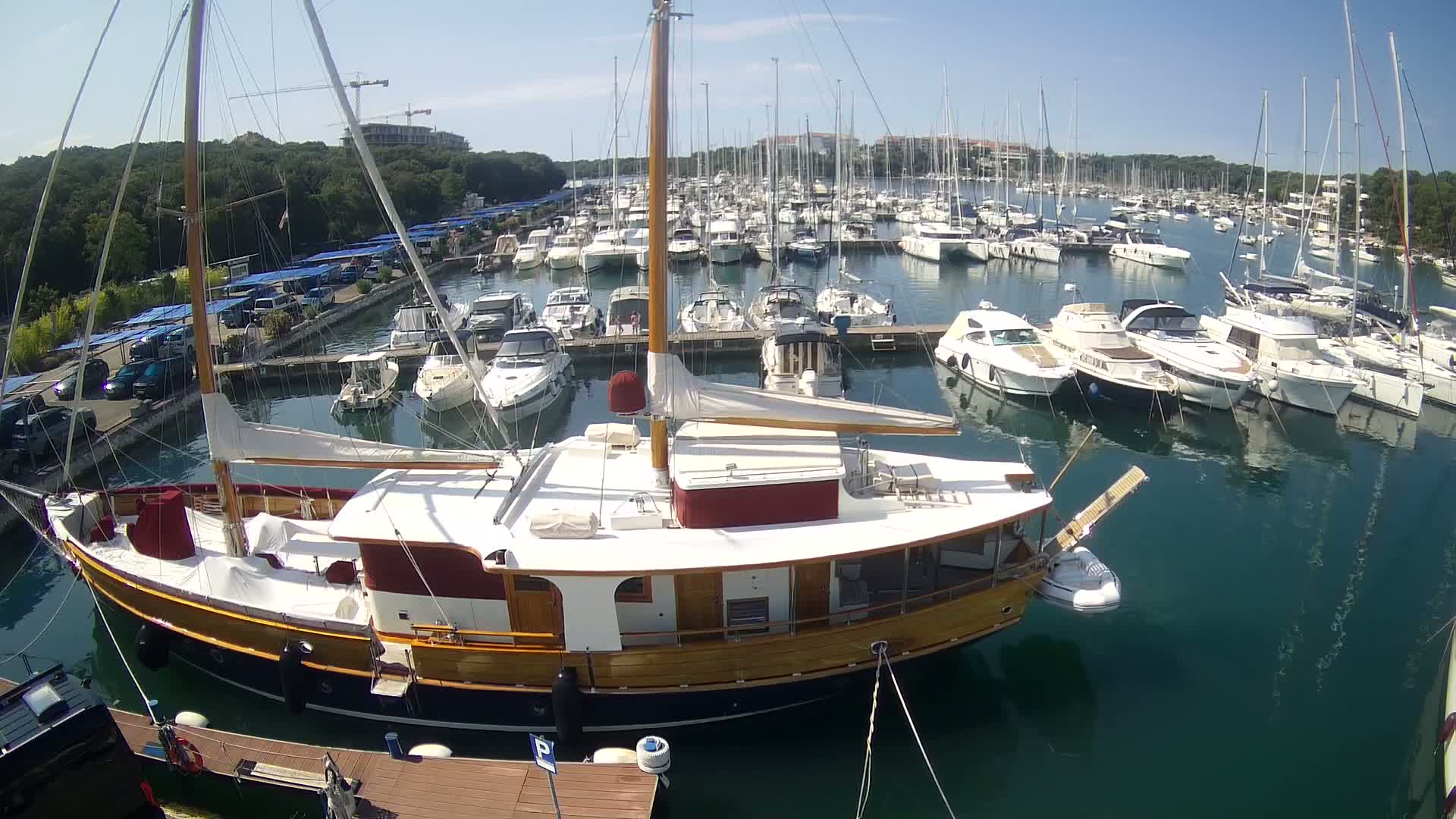 A large wooden sailboat is docked in a marina filled with numerous other boats on a sunny day.