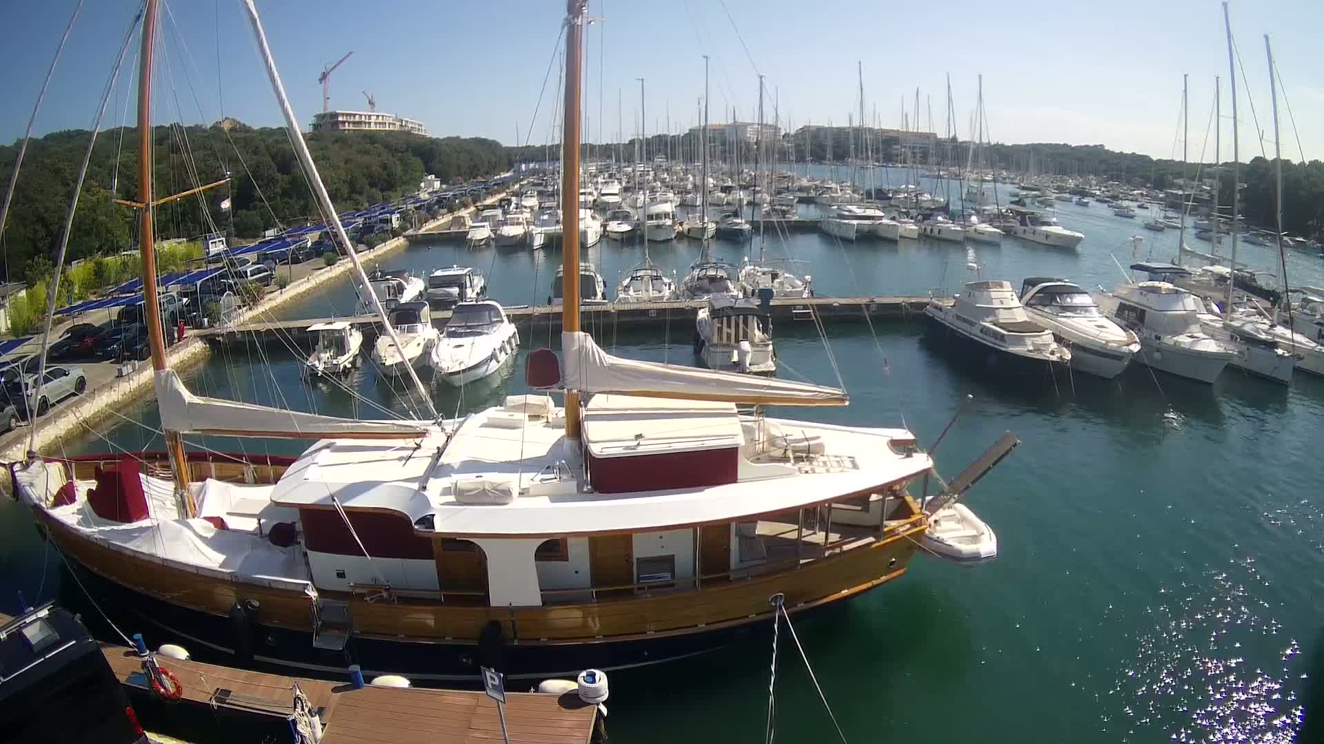 A large wooden sailboat is docked in a marina filled with numerous other boats on a sunny day.