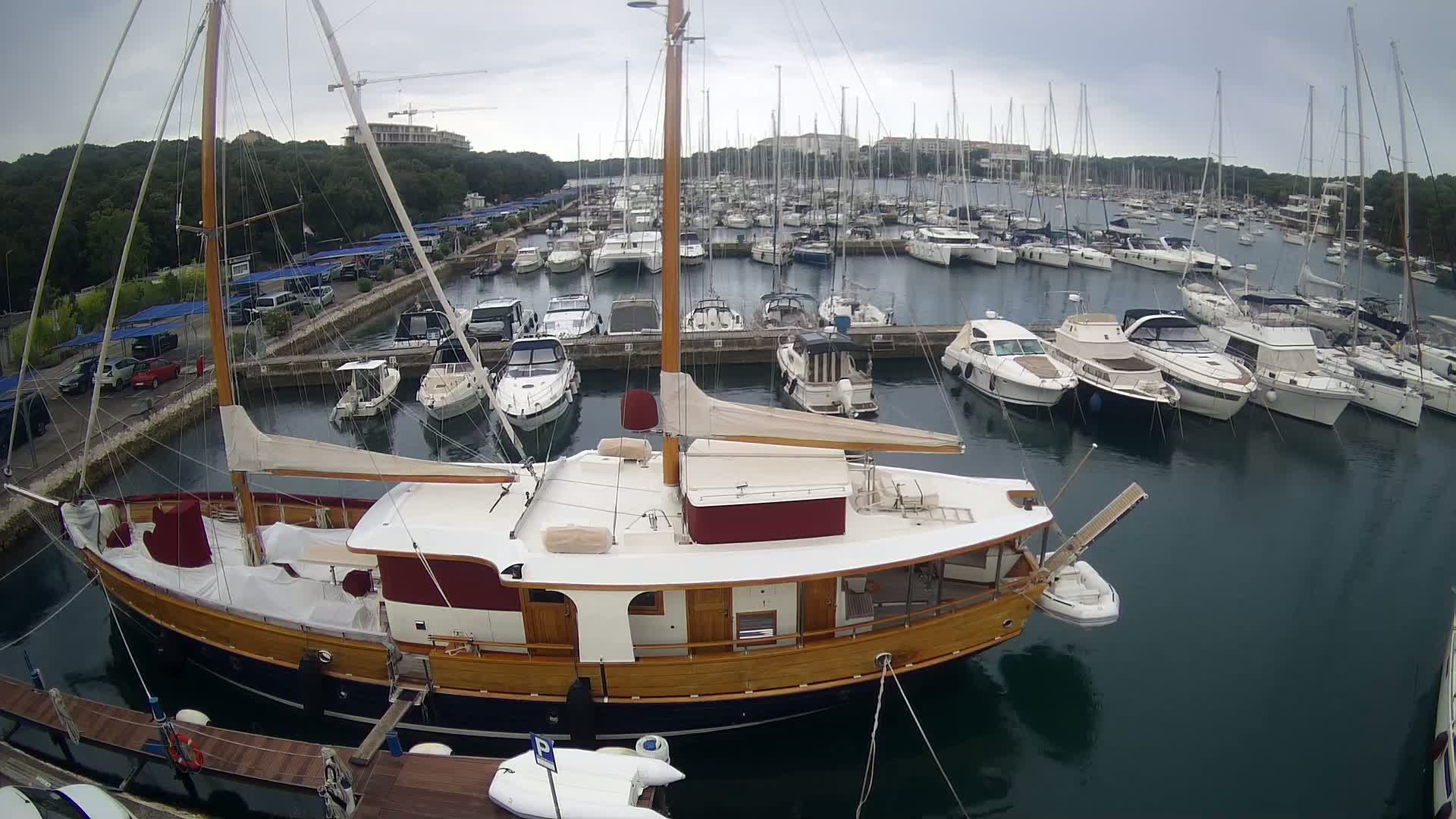 A wooden sailboat is docked in a marina filled with numerous other boats on an overcast day.