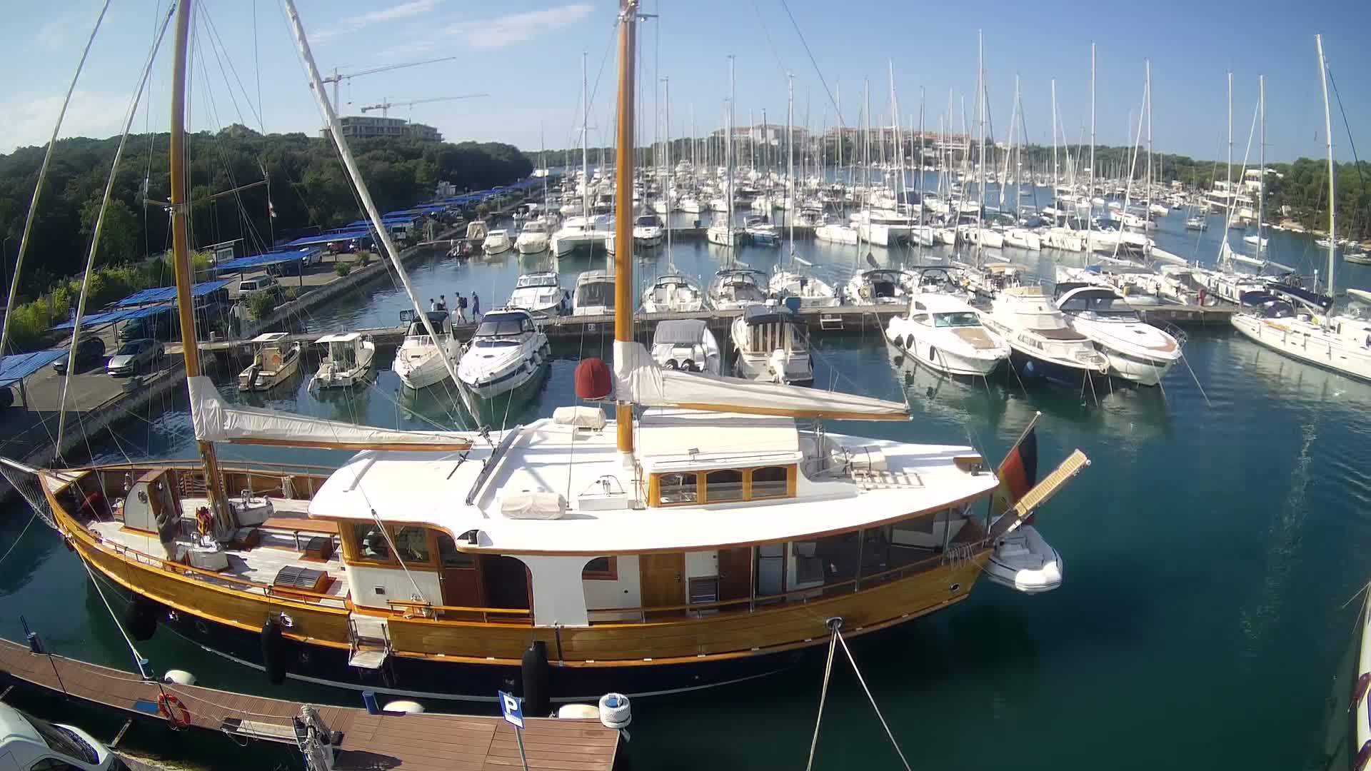 A large, wooden-hulled sailboat is docked in a marina filled with numerous other sailboats and motorboats on a sunny day.