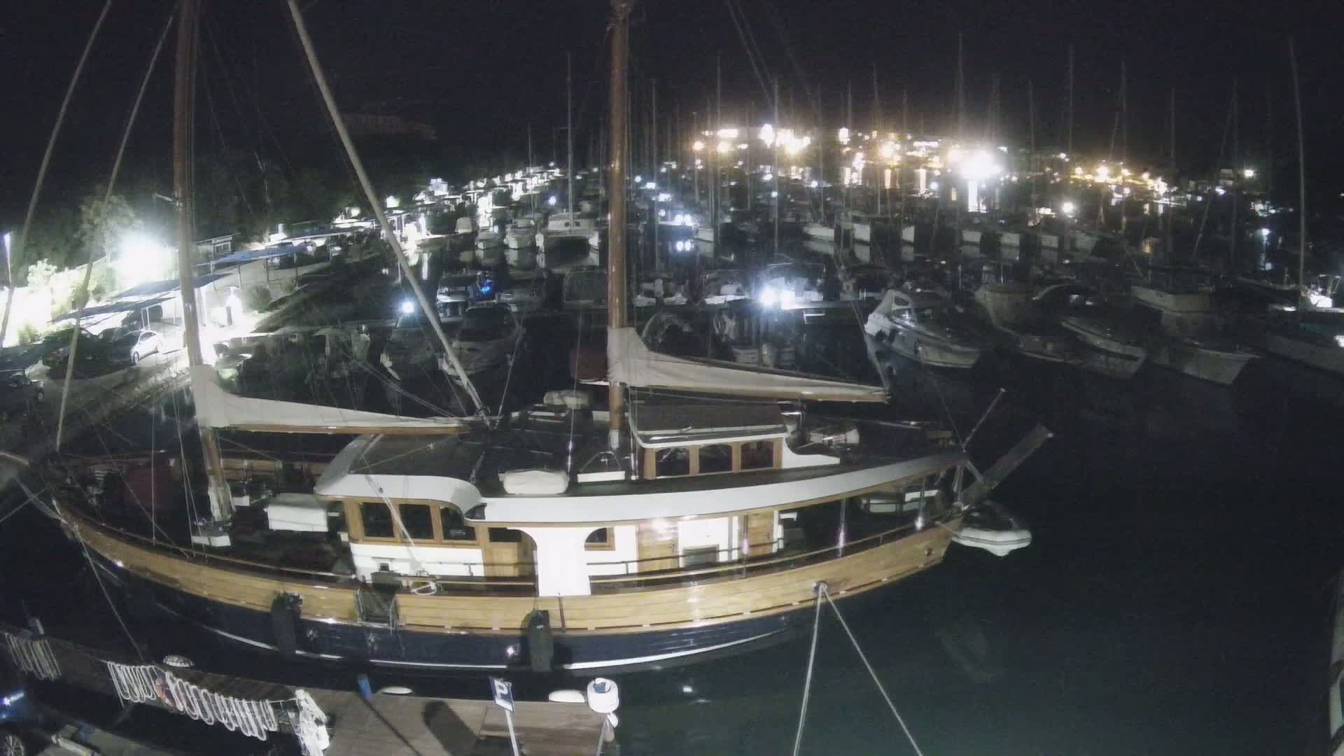 A large, wooden-hulled sailboat is docked in a marina filled with numerous other sailboats and motorboats on a sunny day.