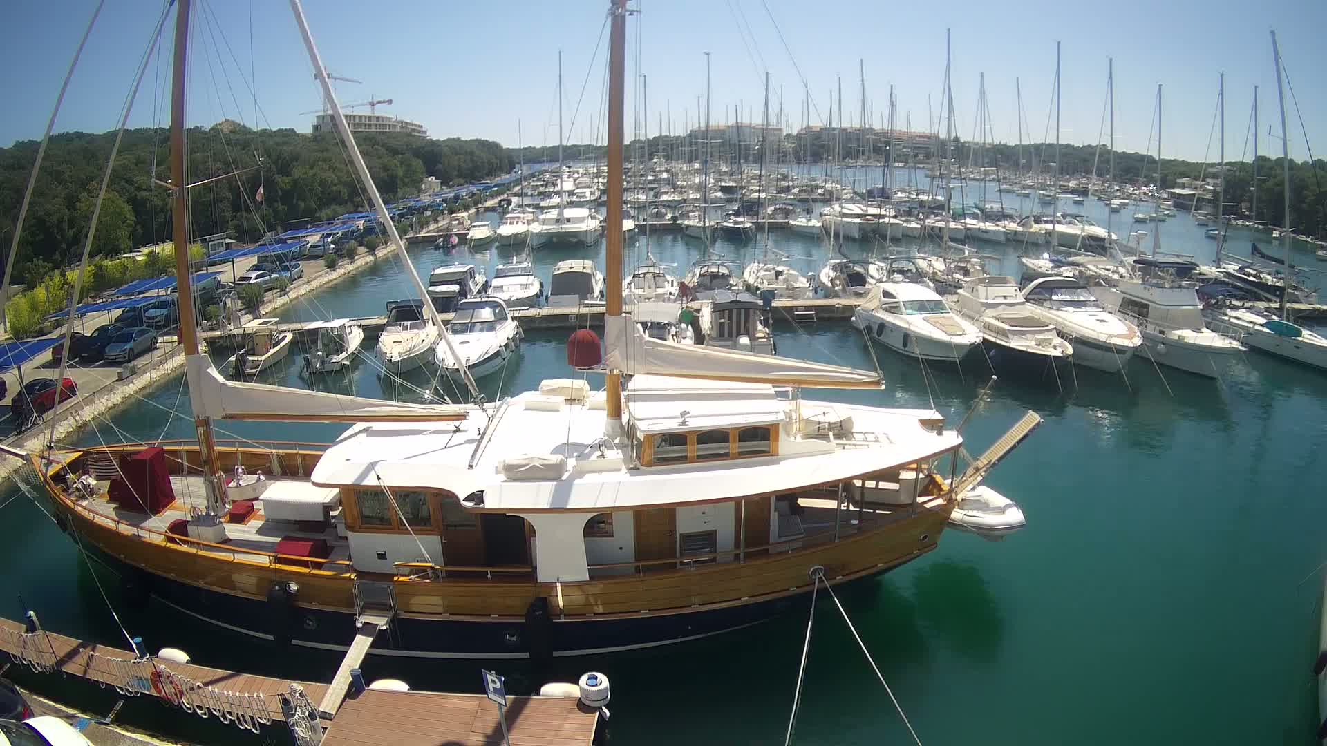 A large, wooden-hulled sailboat is docked in a marina filled with numerous other sailboats and motorboats on a sunny day.