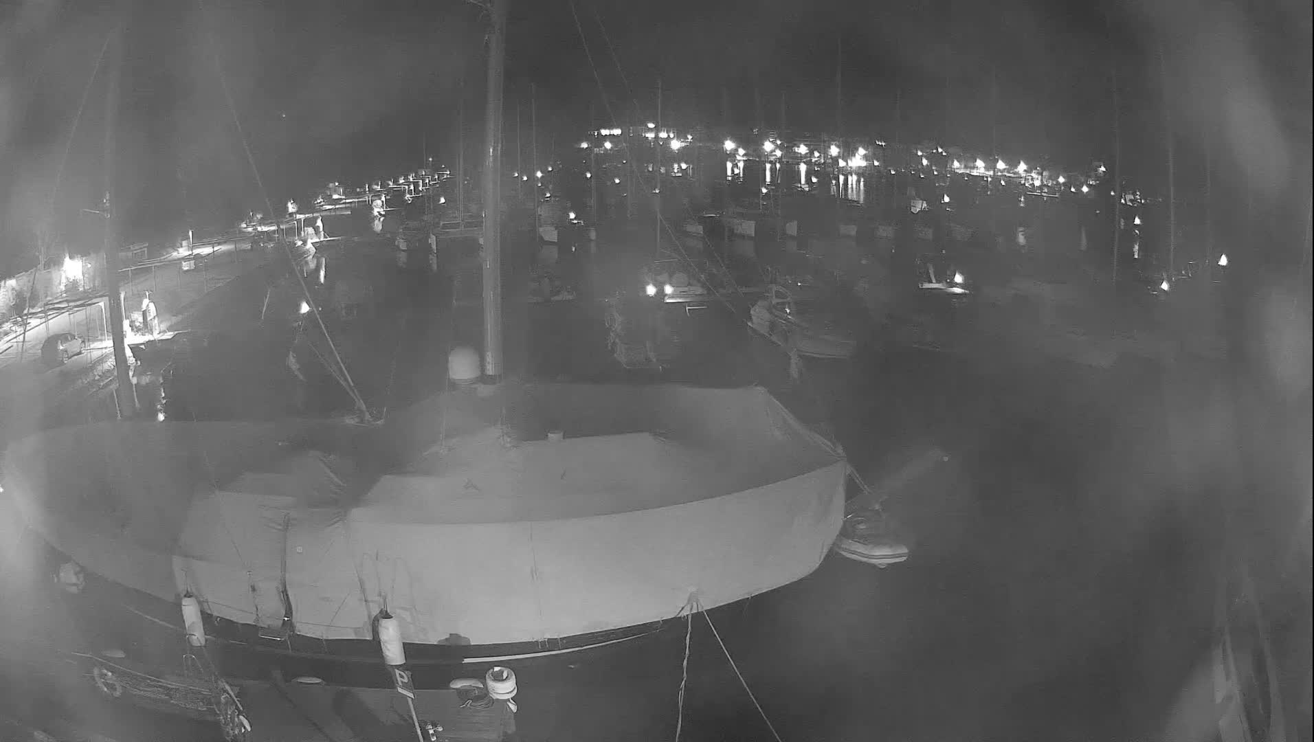 An elevated grayscale image captures a quiet marina at night, filled with numerous docked boats illuminated by pier lights reflecting on the calm water under a clear sky.