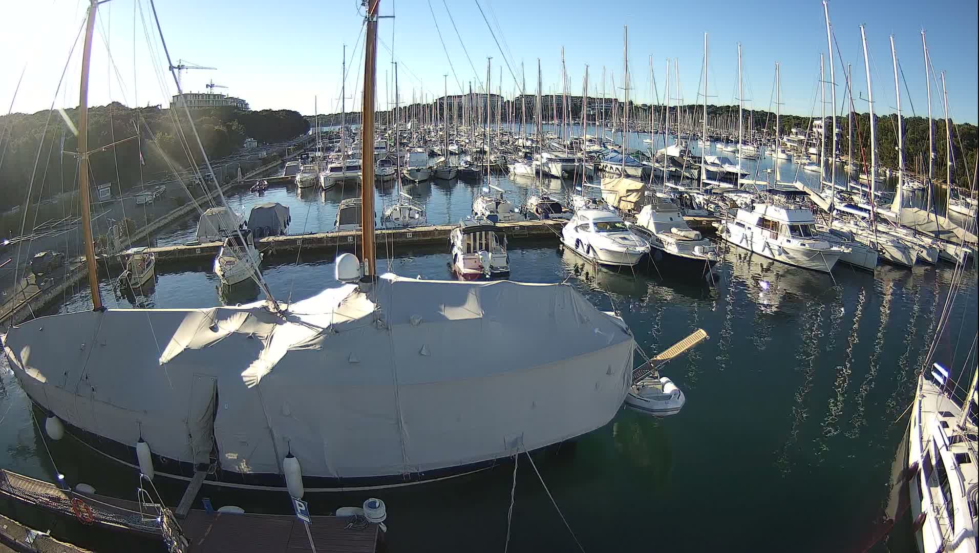 A busy marina is filled with numerous sailboats and yachts under a clear blue sky on a bright, sunny day, with a large covered sailboat prominent in the foreground.