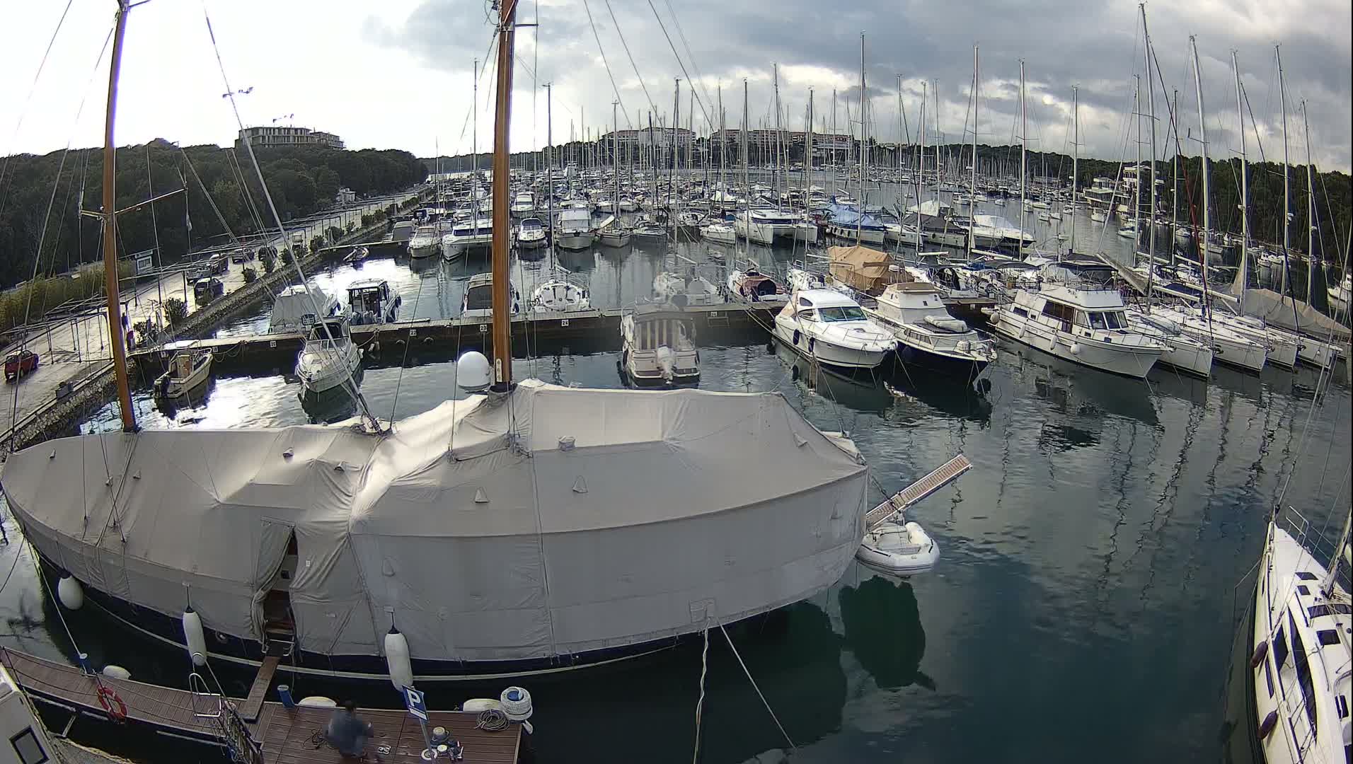 A vast marina filled with numerous sailboats and motor yachts, including a large boat covered with a light grey tarp in the foreground, is observed under a heavily overcast and cloudy sky.