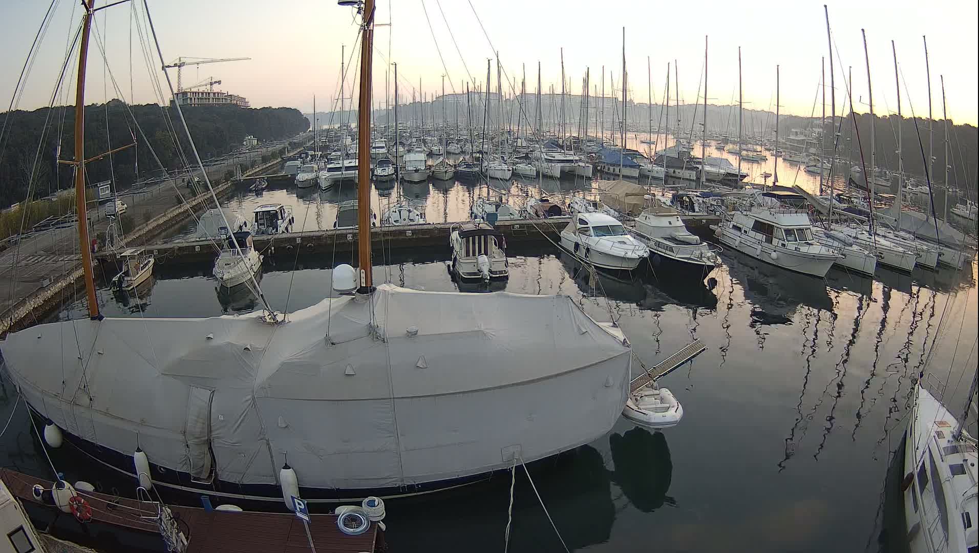A vast marina filled with numerous sailboats and motor yachts, including a large boat covered with a light grey tarp in the foreground, is observed under a heavily overcast and cloudy sky.