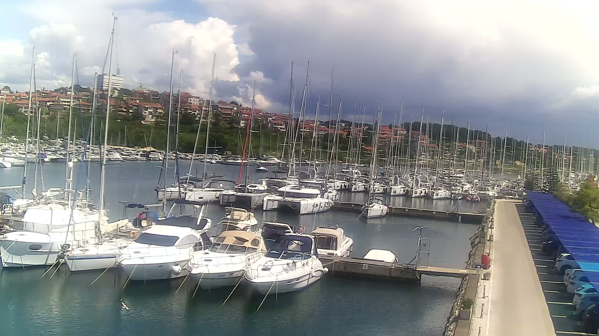 A marina filled with numerous sailboats and motorboats is situated near a hillside of houses under a partly cloudy sky.