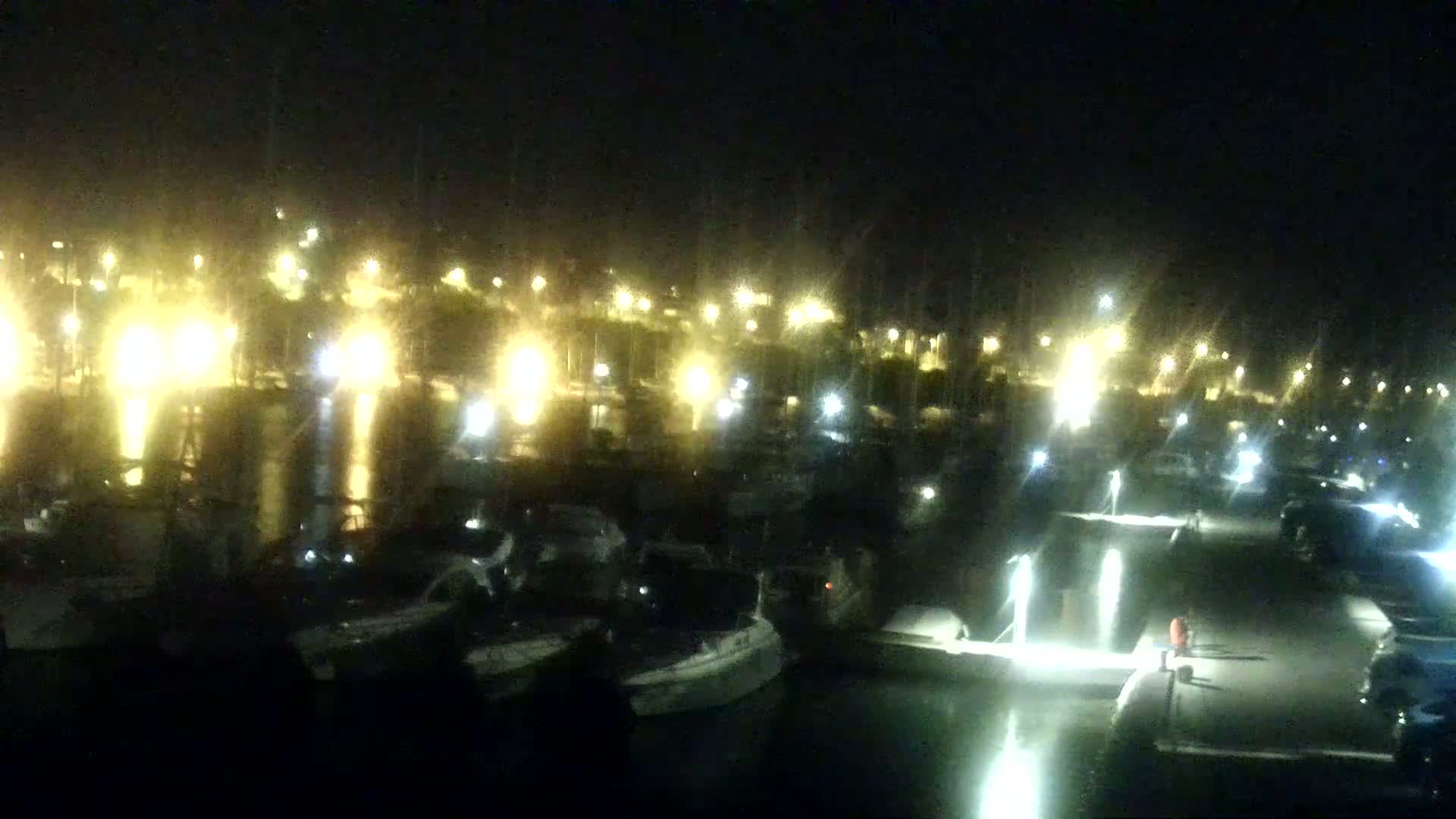 Numerous boats are docked at a marina at night, illuminated by many lights reflecting on the water.