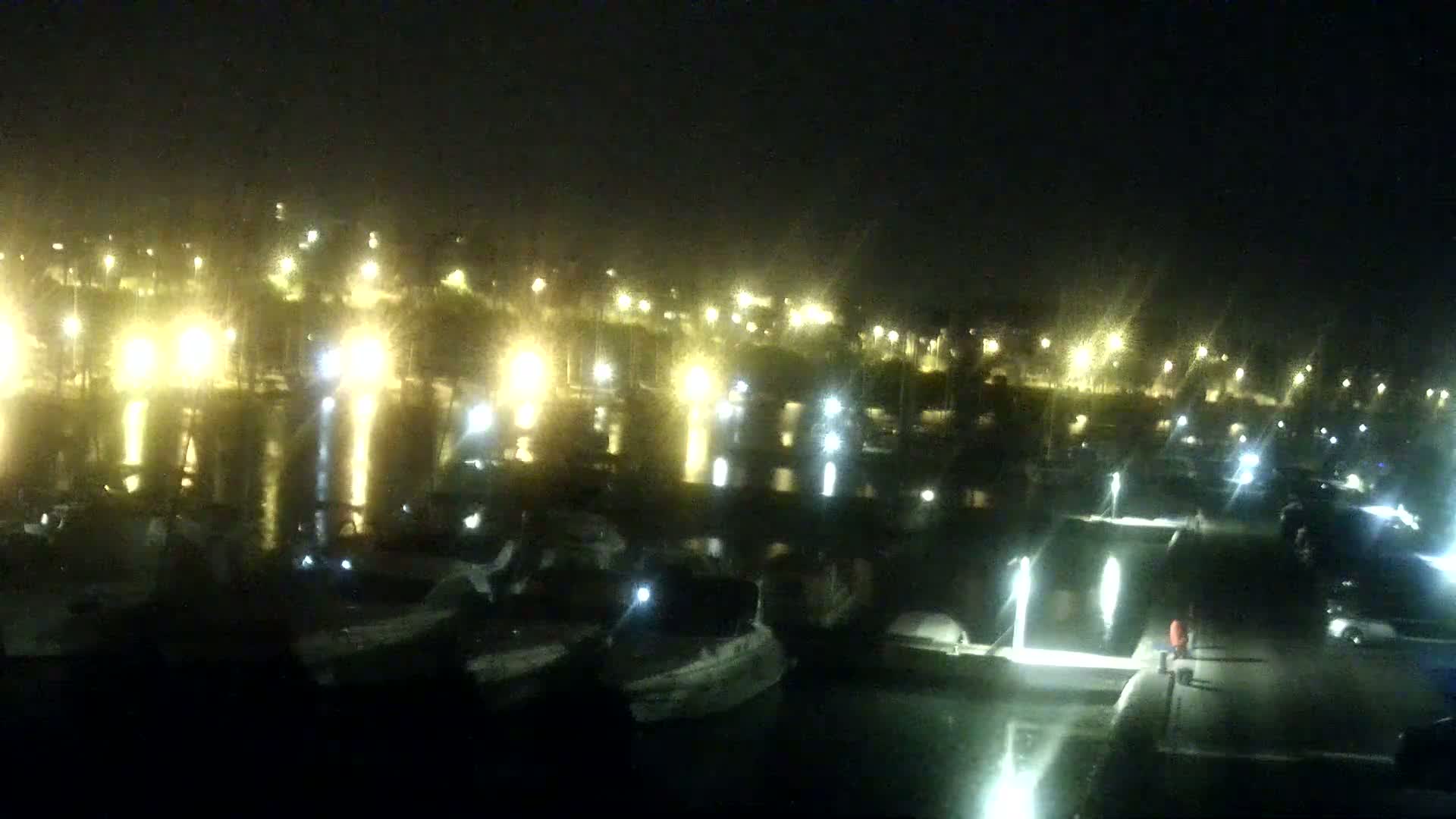 A nighttime view of a marina filled with boats, illuminated by numerous lights reflecting on the calm water.