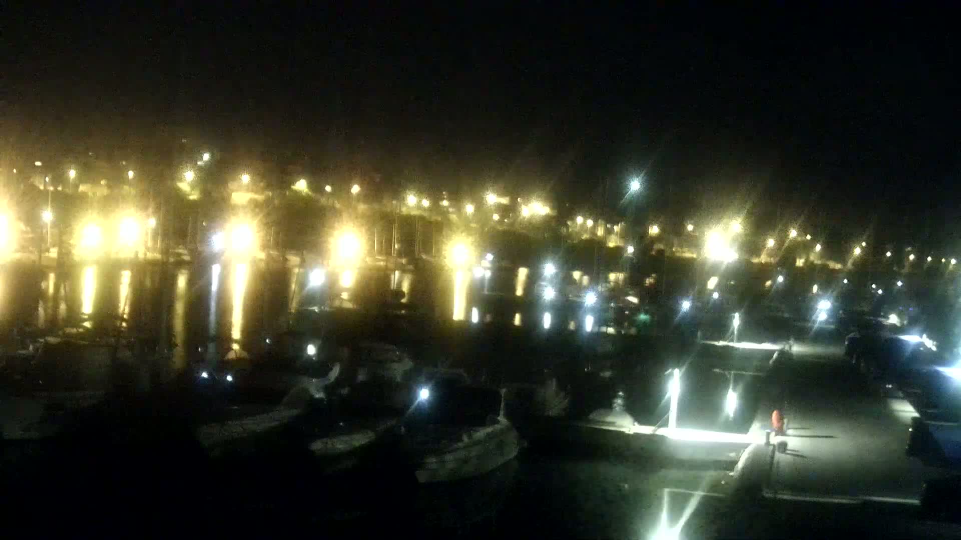 A nighttime view of a marina, illuminated by many lights, reflecting on the calm water.