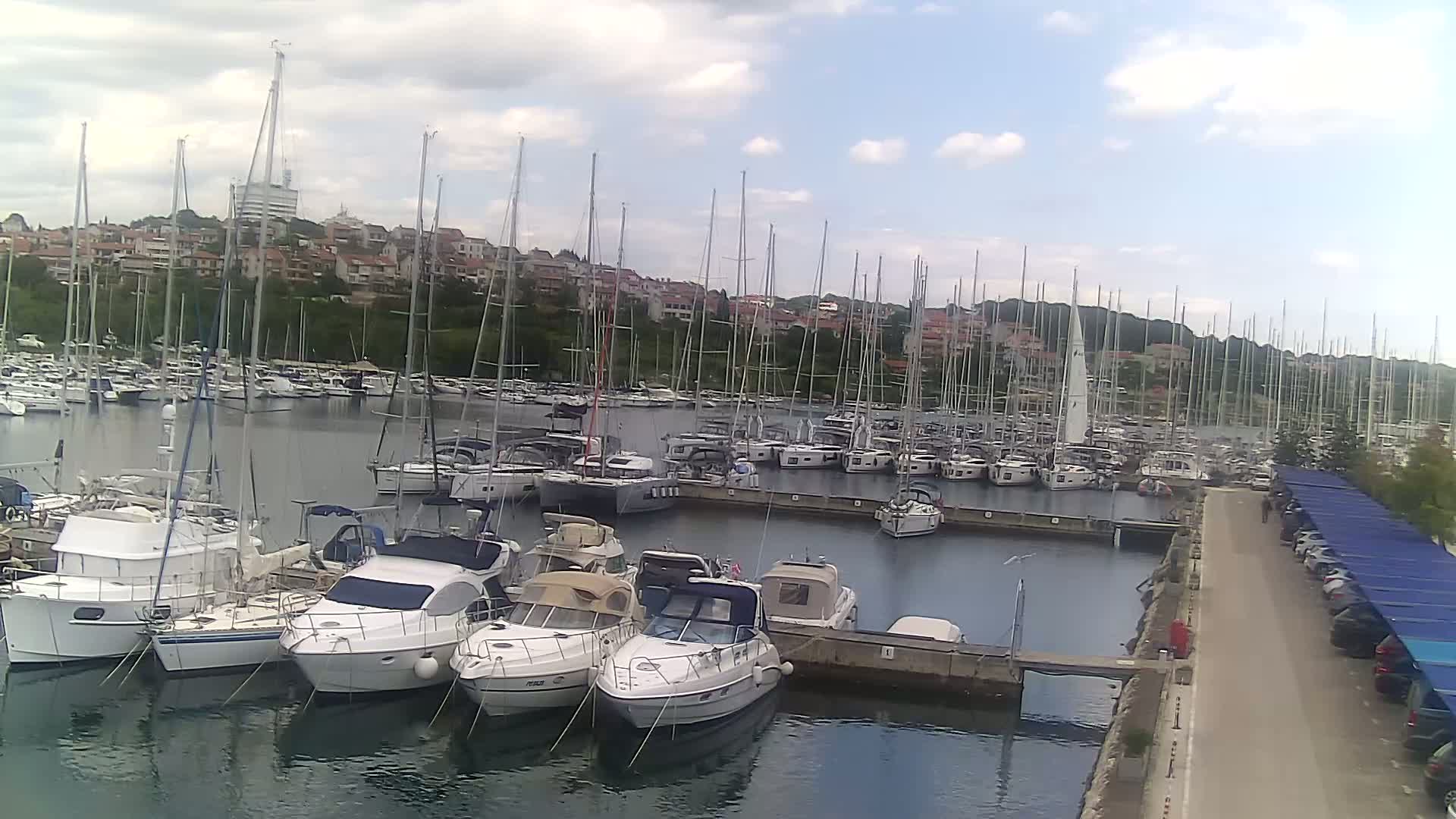 A marina filled with numerous sailboats and motorboats is situated on calm water under a partly cloudy sky.