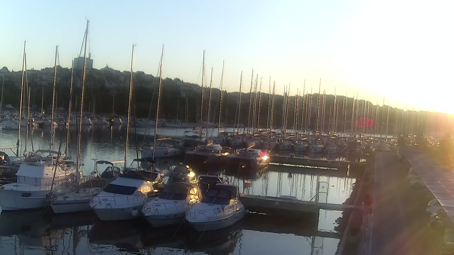 Many sailboats and motorboats are docked in a marina at sunset, with calm water reflecting the light.
