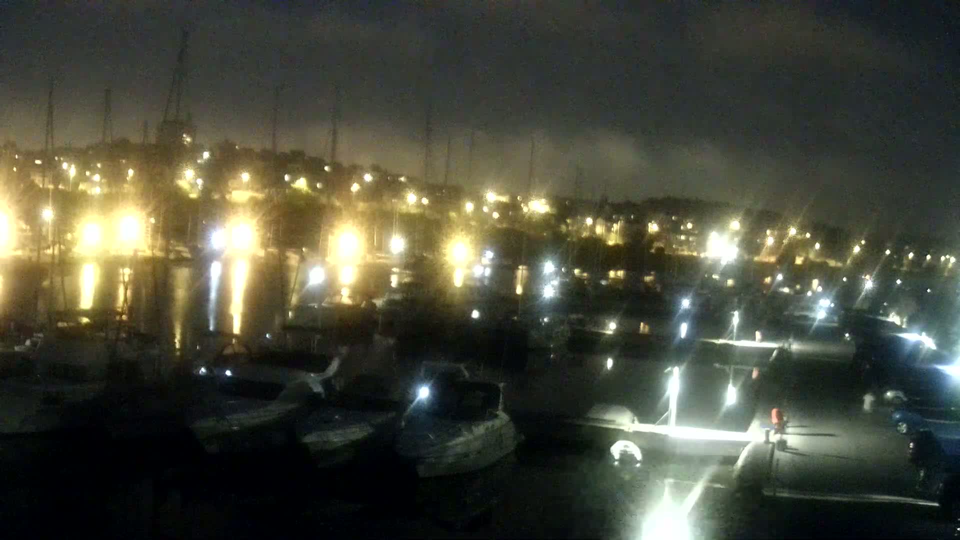 A nighttime view of a marina with many boats docked, illuminated by lights, under a dark and slightly hazy sky.