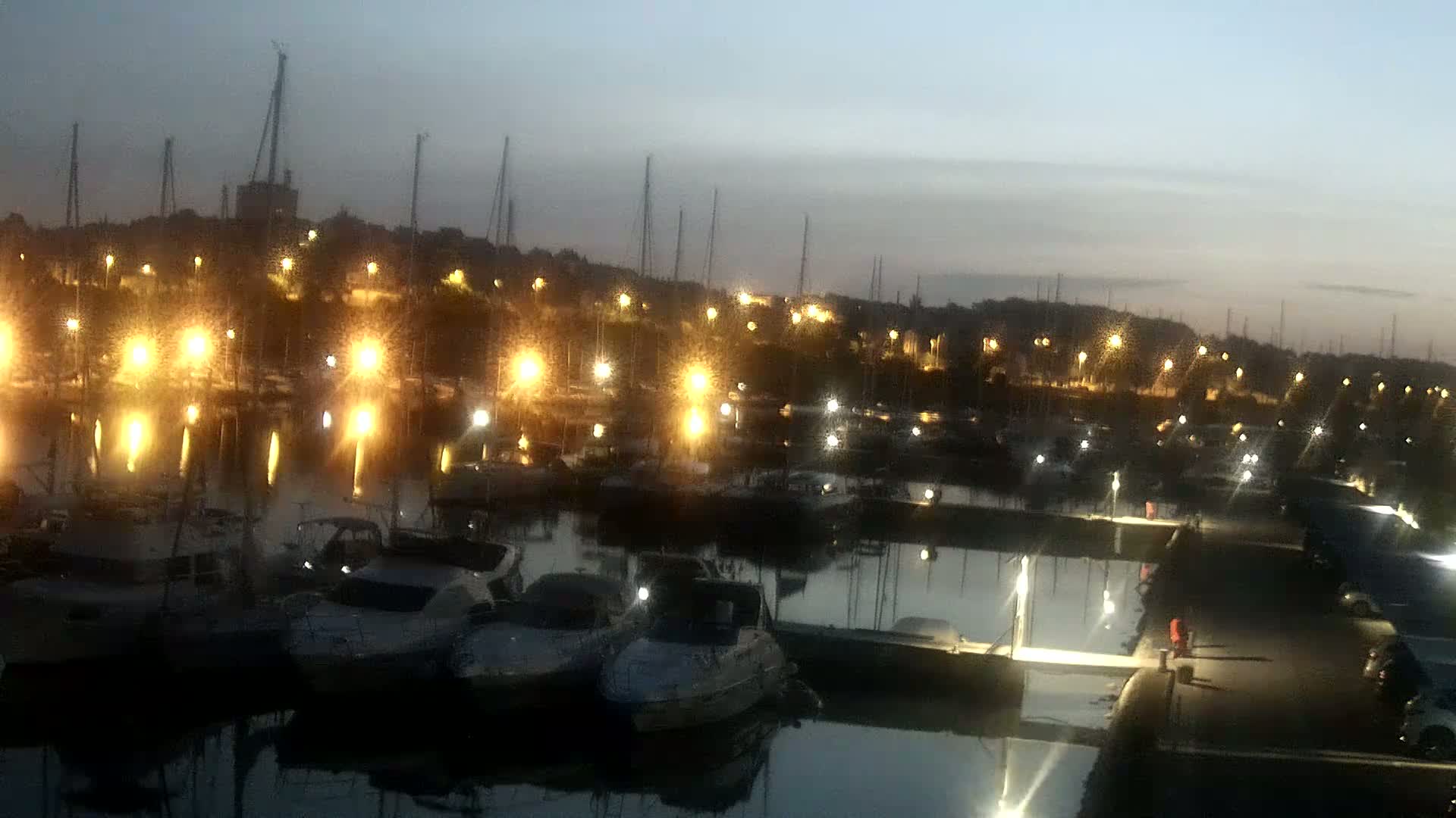 Many boats are docked at a marina at twilight, illuminated by numerous lights reflecting on the calm water.