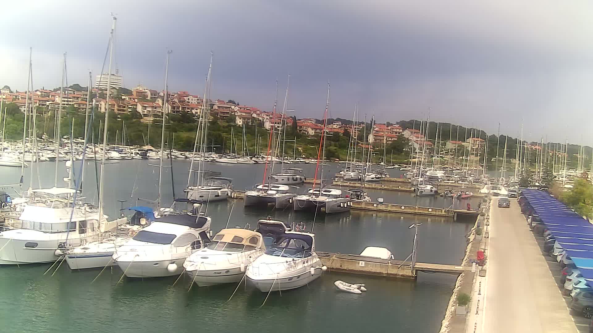 A marina filled with numerous sailboats and motorboats is situated under an overcast sky, with a residential area visible in the background.